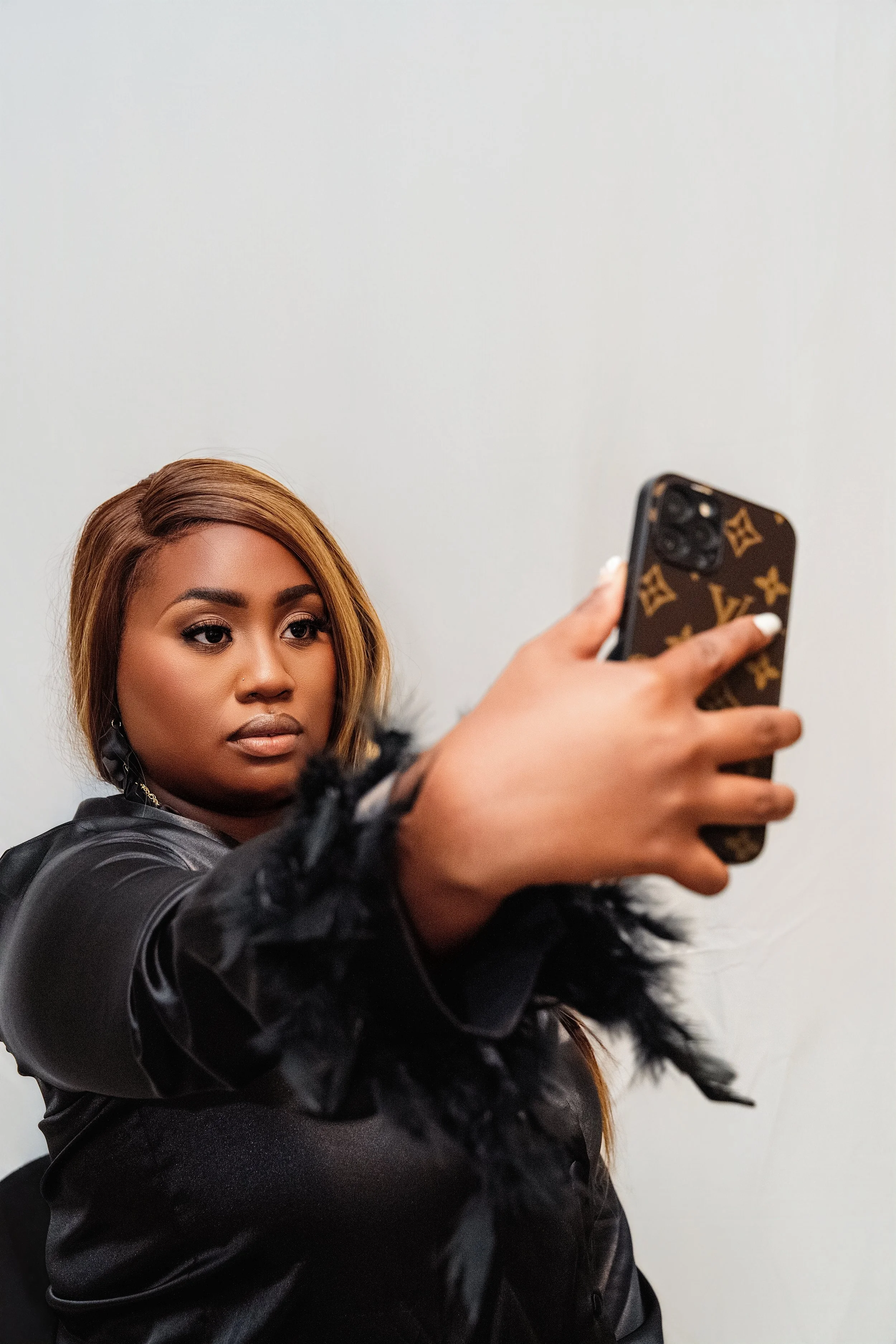 A woman with long light brown hair taking a selfie with her smartphone, wearing a black satin jacket with black fur cuffs, standing against a plain white wall.