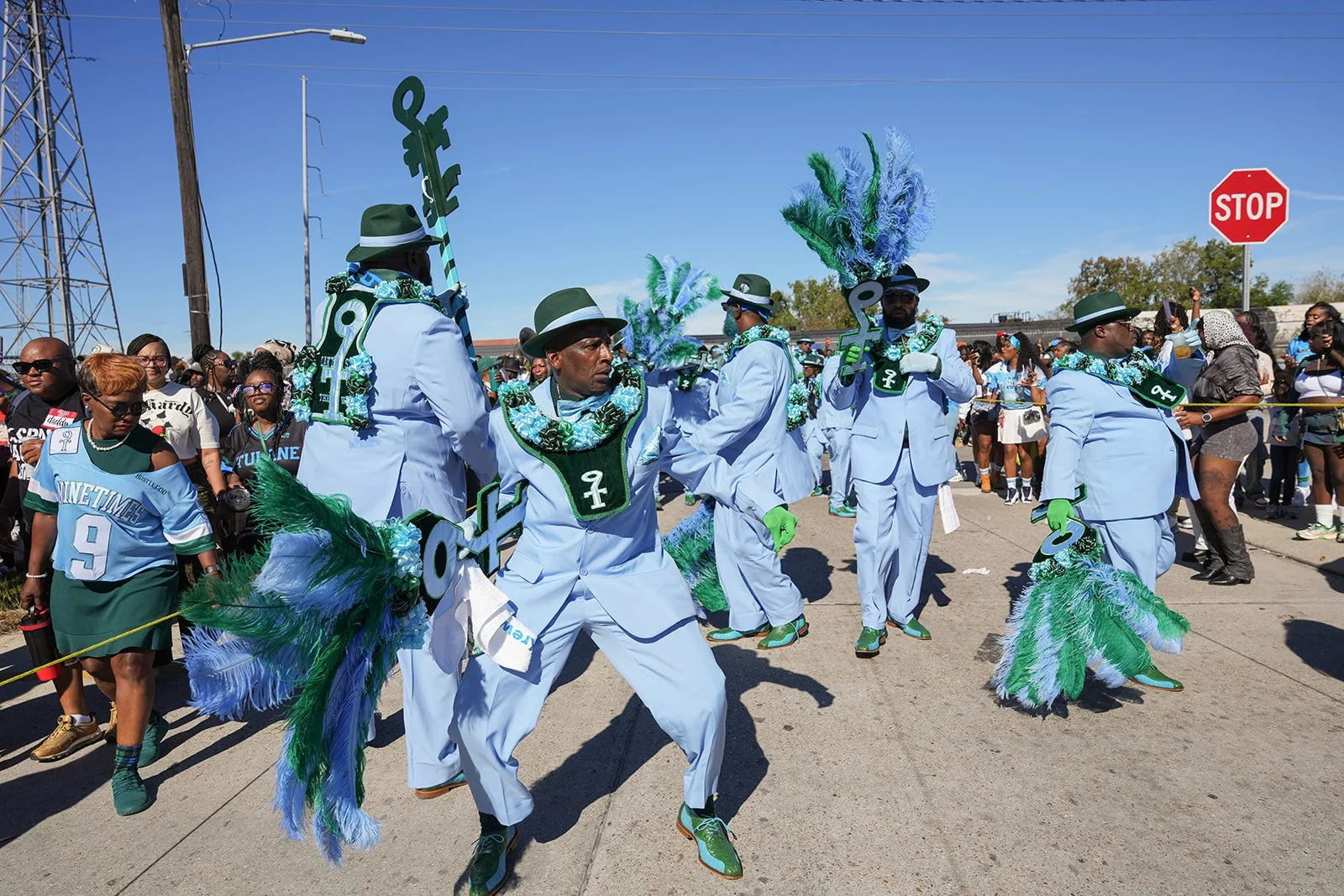 People participating in a parade wearing costumes with green, blue, and white feathers, large keys, and fashionable accessories on a sunny street with onlookers and a stop sign.
