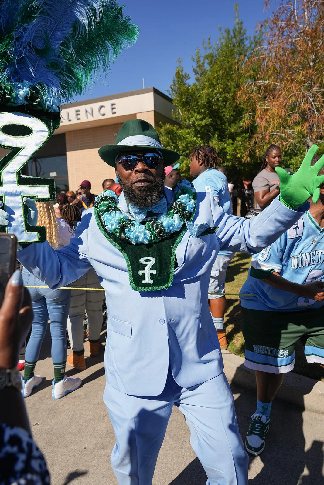 Man wearing a suit and tie, large green and blue flower necklace, green gloves, sunglasses, and a hat, participating in a parade with other people in sports jerseys and casual clothing, holding a sign with the number 2.