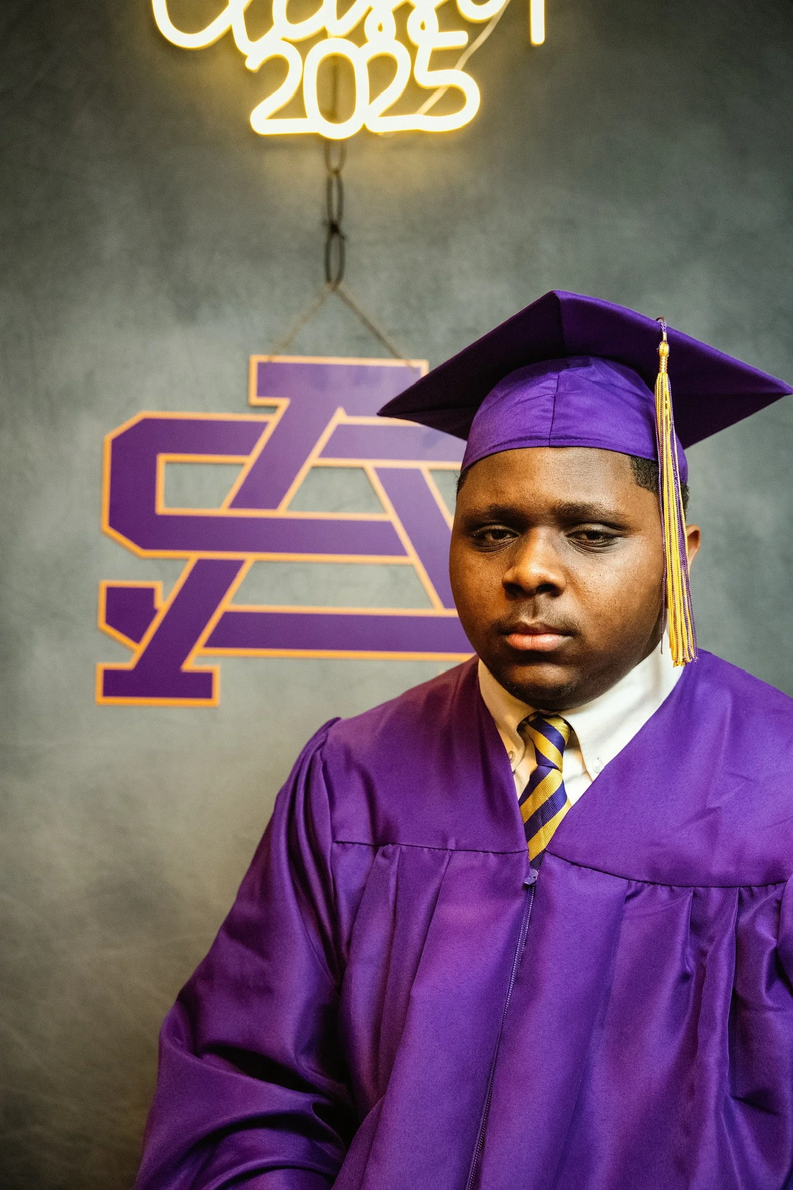 A young man in a purple graduation gown and cap stands indoors, looking down with a serious expression. Behind him is a gray wall with a purple and gold logo and a neon sign saying 'Class of 2023'.