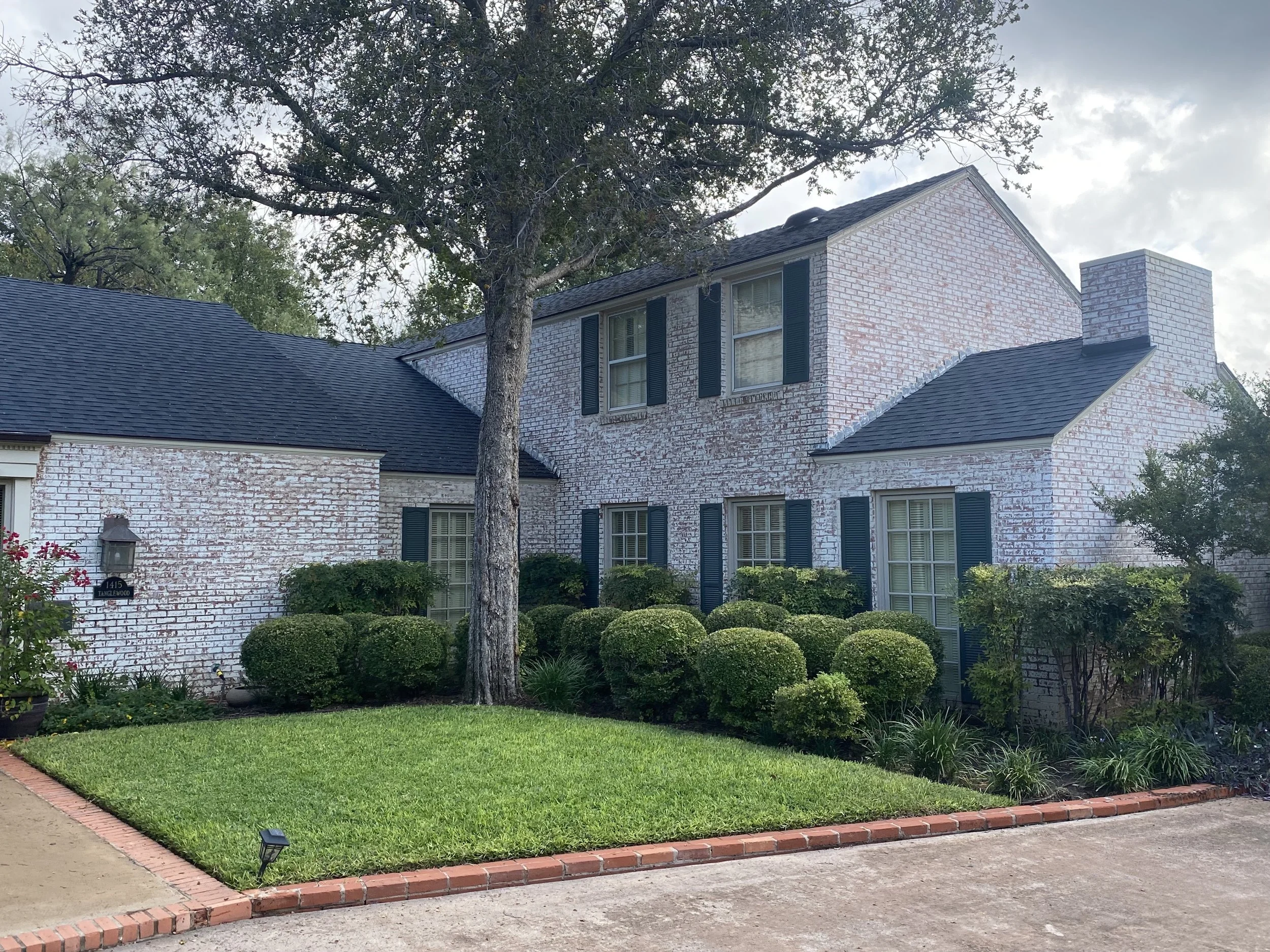 Two-story brick house with blue shutters, black roof, front yard with green grass, bushes, and trees, on a cloudy day.