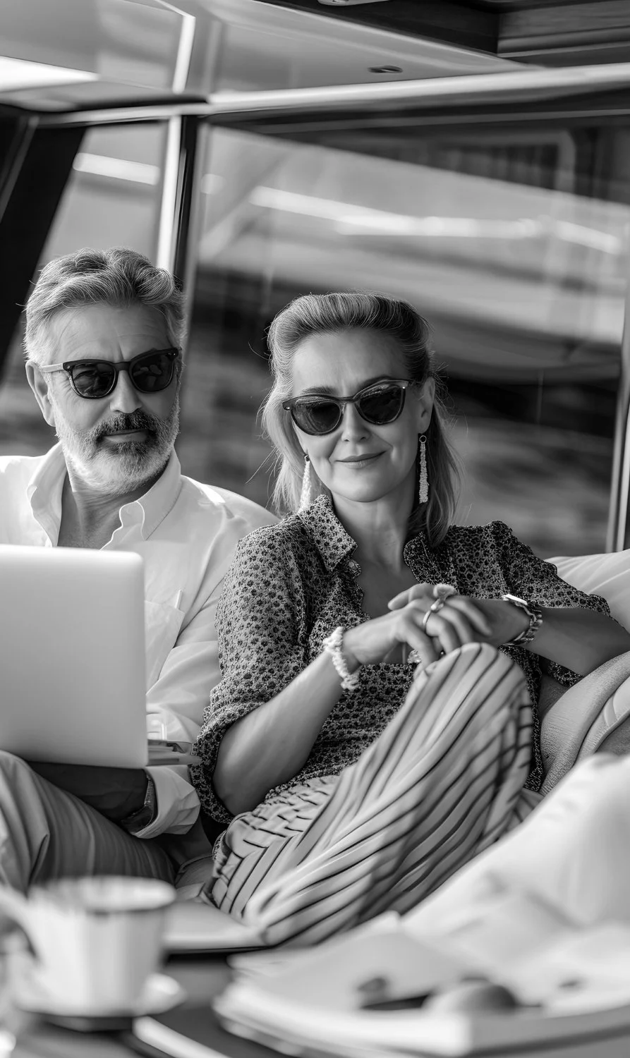 A black and white photo of a mature man and woman sitting together with sunglasses, looking at a laptop, outside on a sunny day.