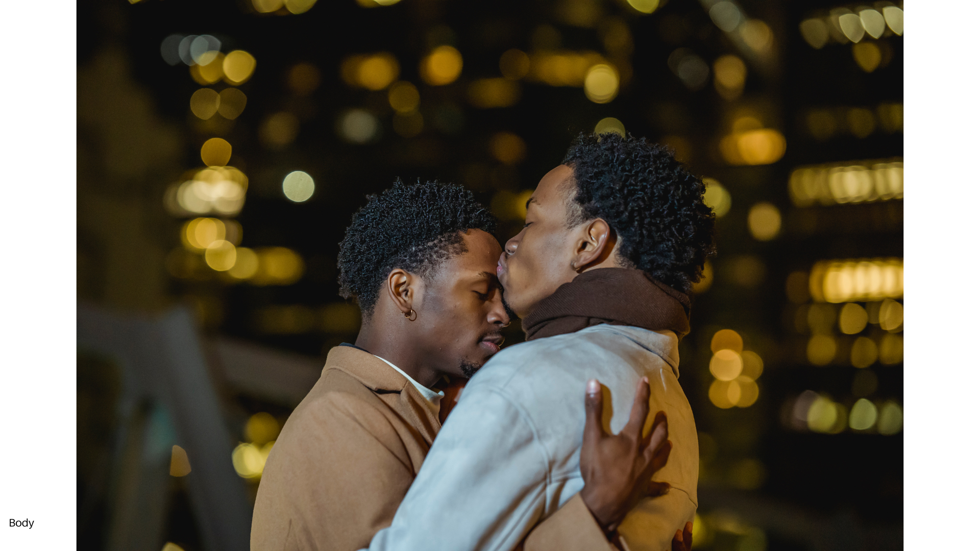 Two men embrace closely, touching foreheads with eyes closed against a blurred city lights background at night.