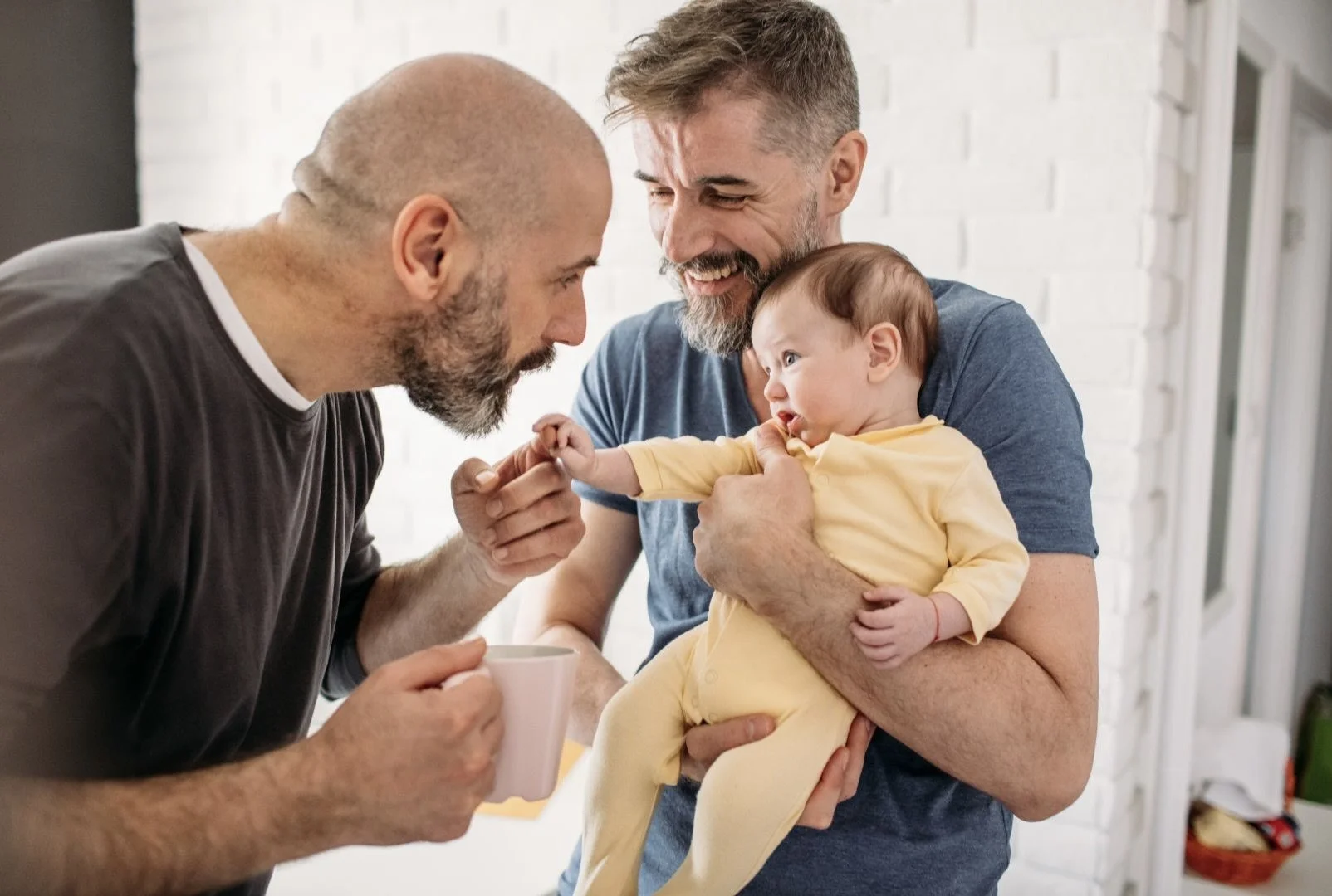 Two adult men and a baby are smiling and interacting closely. One man is holding the baby, who is reaching out and touching the face of the other man.