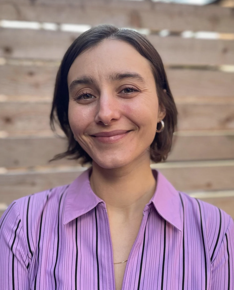 A young woman with short dark hair, light skin, and a septum piercing, smiling outdoors in front of a wooden background, wearing a purple striped shirt.