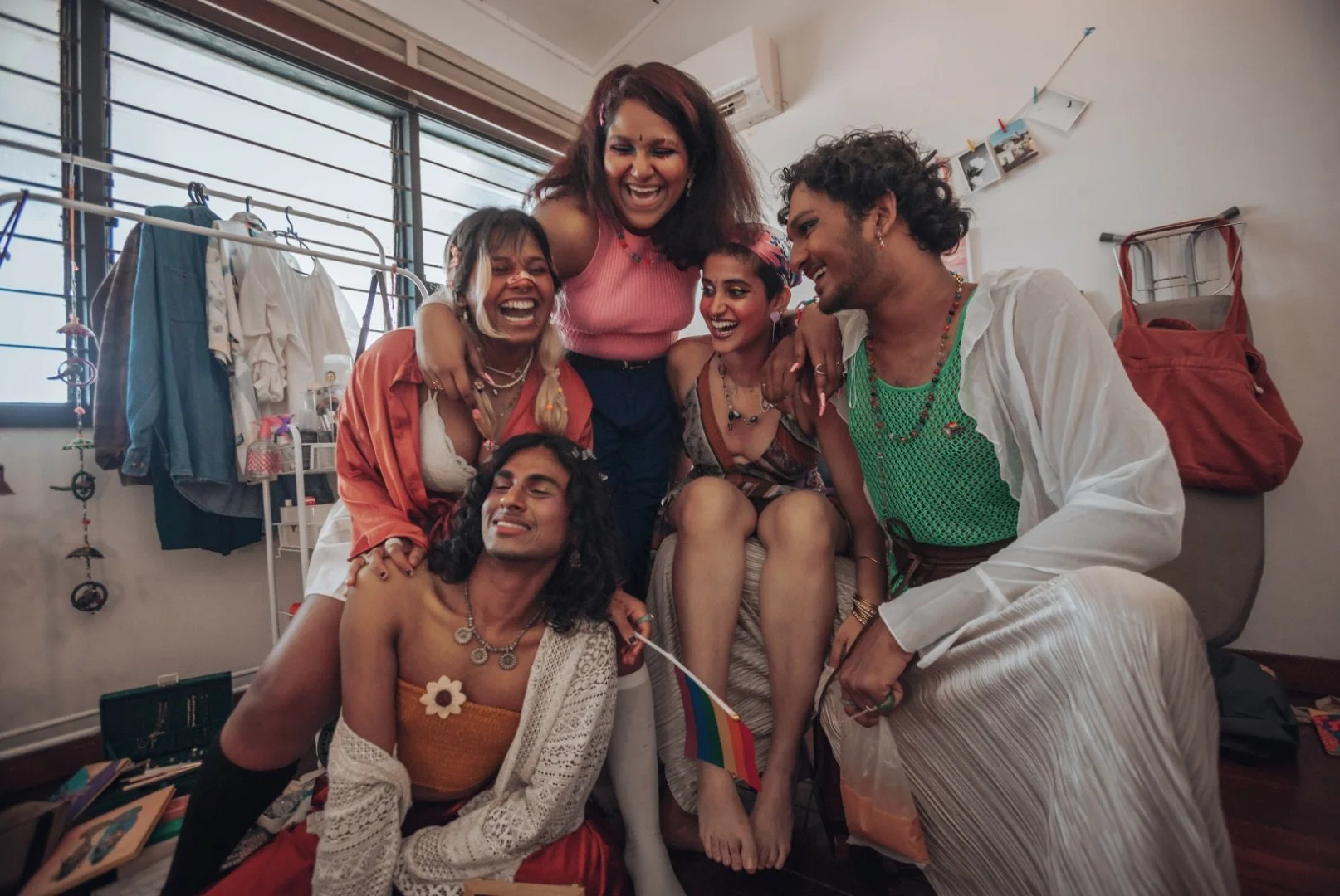 Group of six diverse friends joyfully laughing and hugging indoors, surrounded by personal items and laundry, celebrating Pride with rainbow accessories.