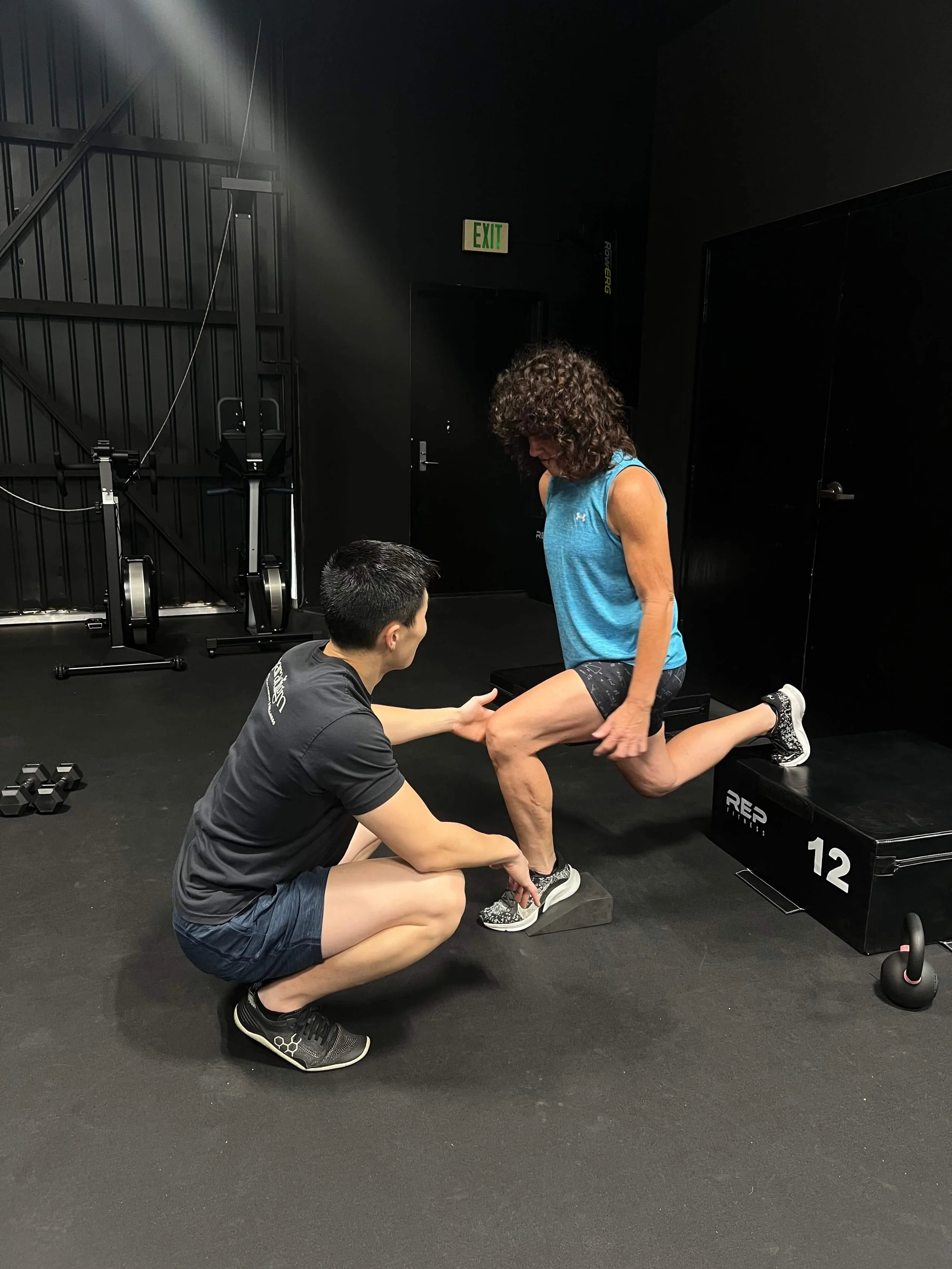 A middle-aged female athlete performing a split squat with her front foot on a wedge. A physical therapist is cueing her to make sure her knee is not caving in.