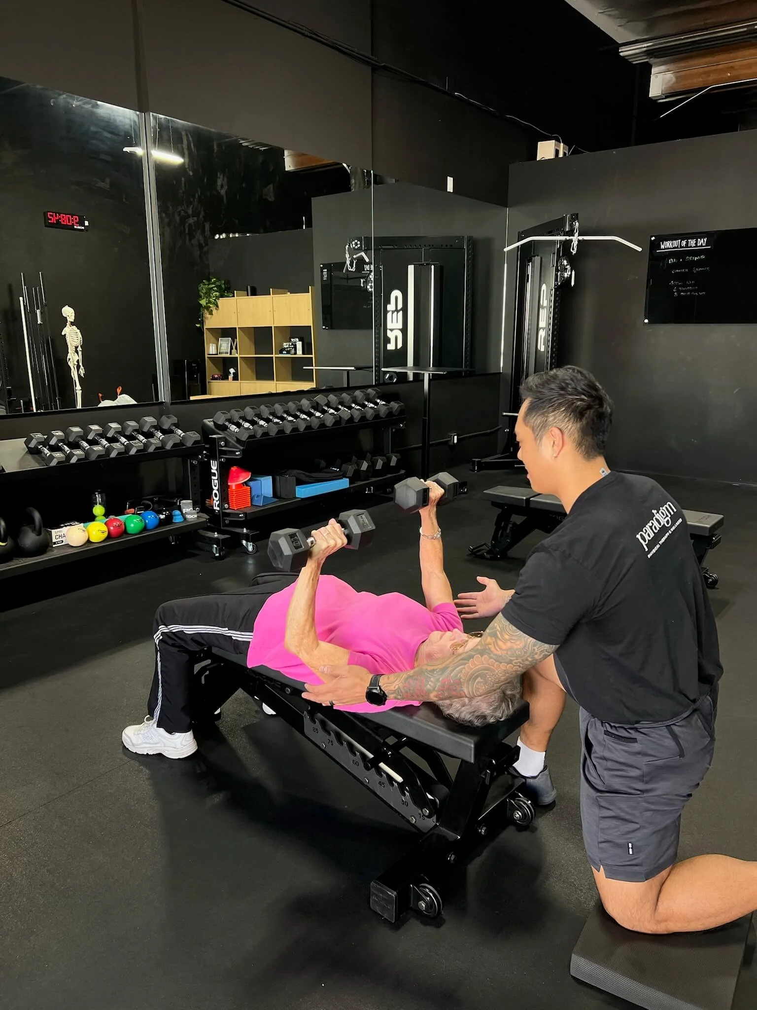 An elderly female performing dumbbell bench presses during her physical therapy session with a physical therapist helping assist during the exercise.