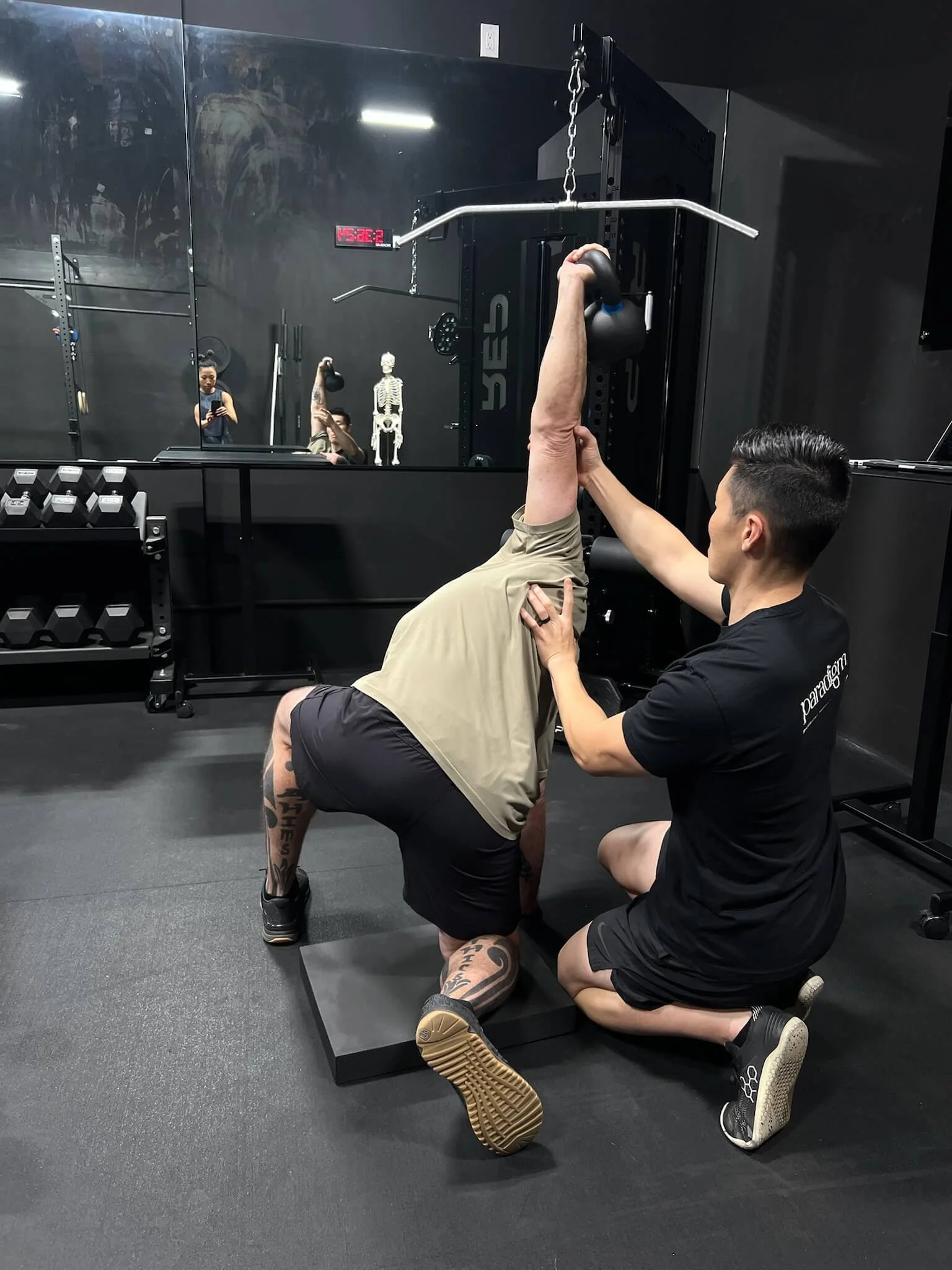 A middle-aged male performing a half-kneeling kettlebell windmill exercise for his shoulder pain while the physical therapist is cueing his scapular motion.