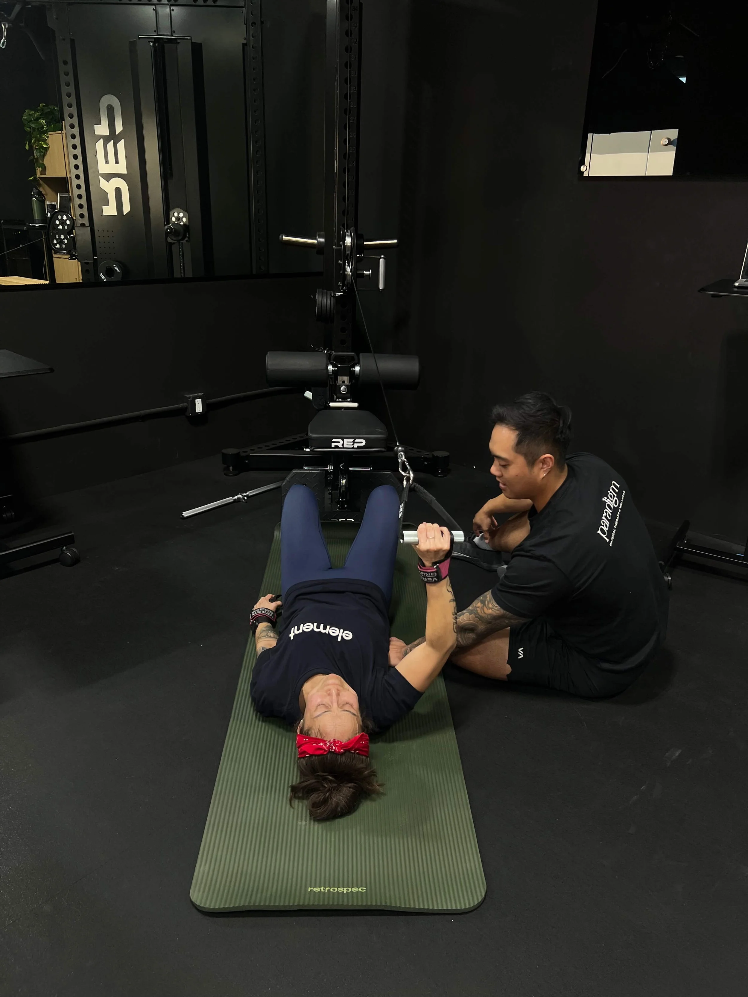 A middle-aged female crossfit athlete laying on the ground performing a single arm cable pull over for her low back and scoliosis physical therapy program. A doctor of physical therapy at is seated beside her for tactile cueing during the exercise.