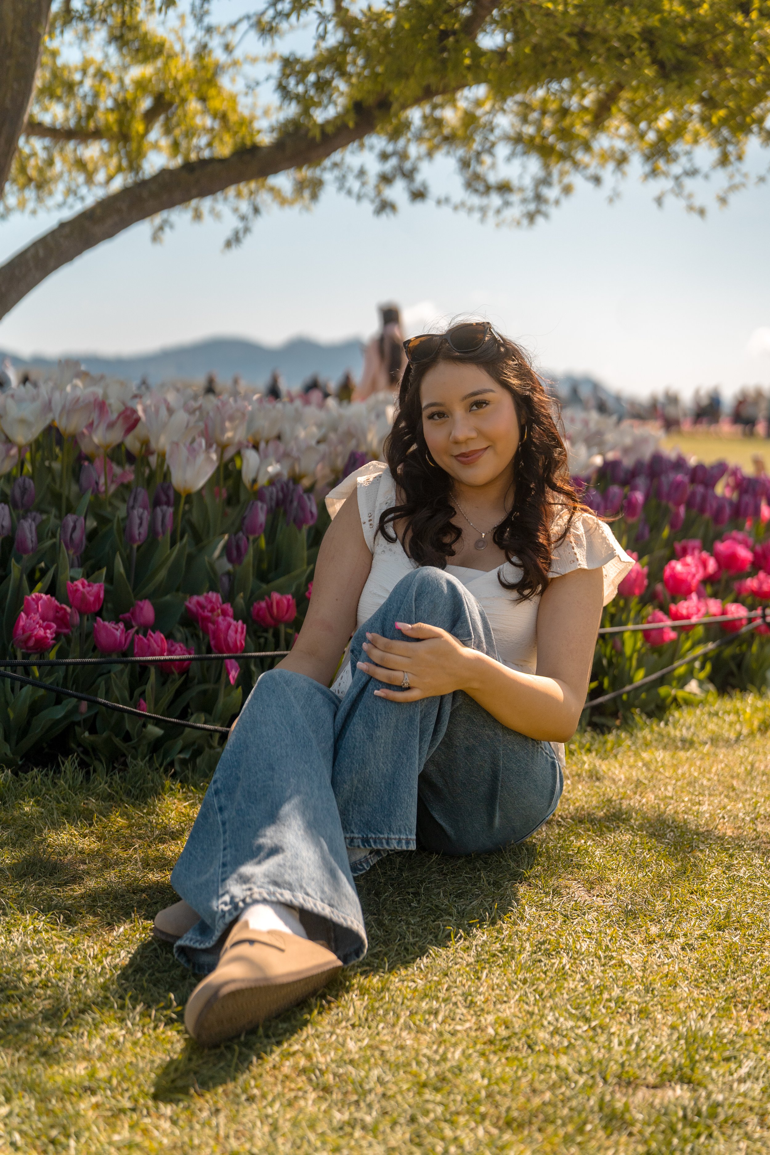 A woman sitting on grass near colorful tulips under a tree with mountains in the background.