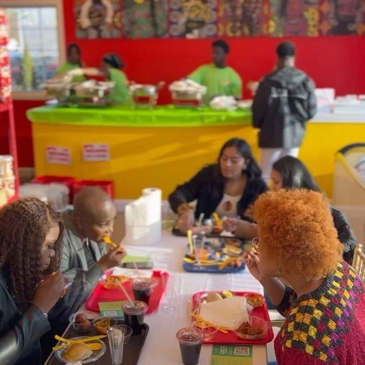 Group of diverse women eating at a table in a colorful restaurant, with a food counter and staff in green uniforms in the background.