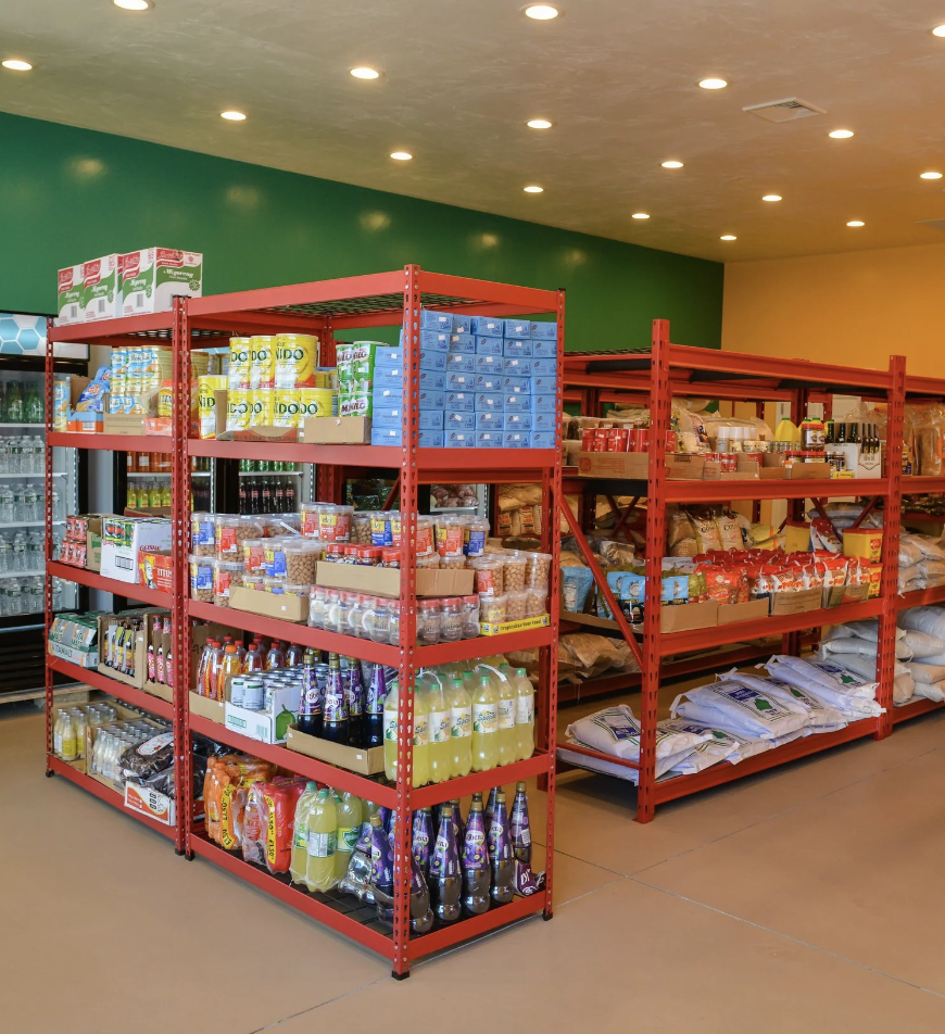 Red metal shelves stocked with food and drink supplies in a store aisle, with a green and yellow wall and ceiling lights overhead.