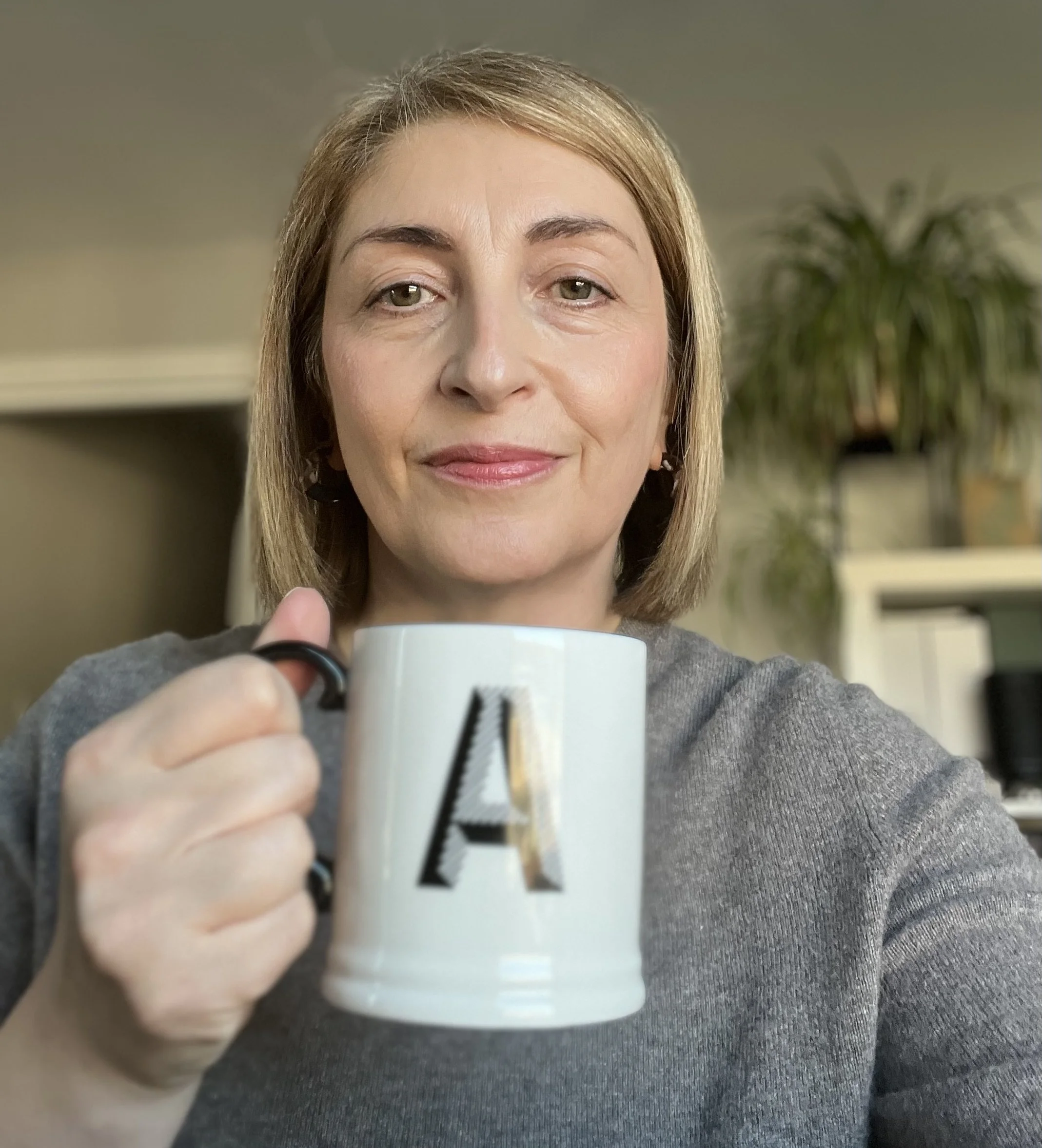 A woman with blond hair holding a white mug with a black letter A, smiling slightly, in an indoor setting with a plant and shelves in the background.
