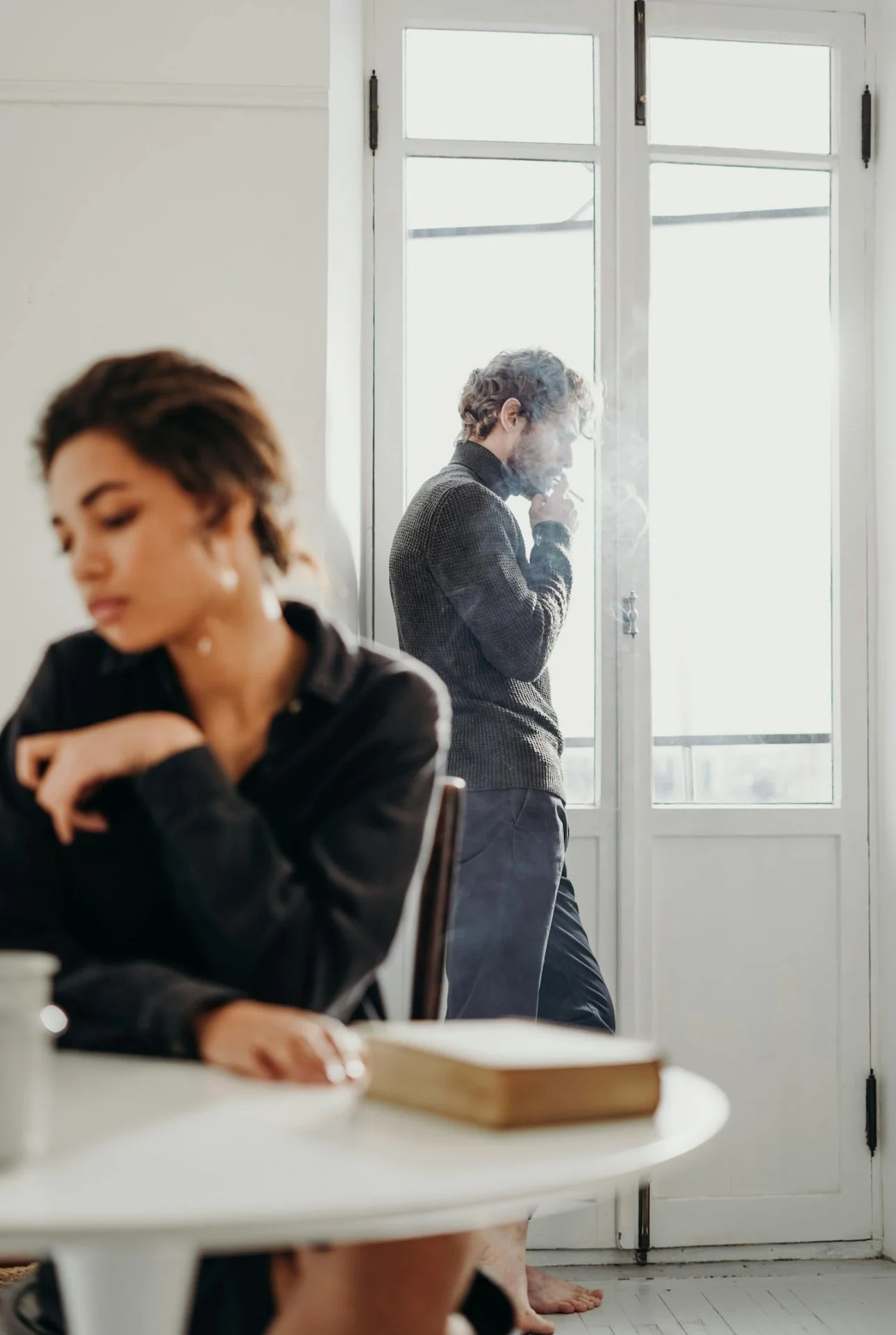 A woman sitting with her eyes closed at a table, while a man in the background is standing by a door, smoking a cigarette, with sunlight coming through the window.