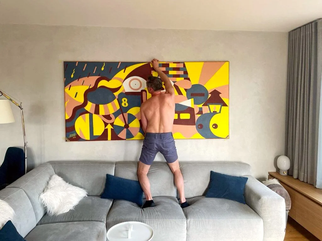 Shirtless man hanging colorful abstract painting on wall above gray couch.