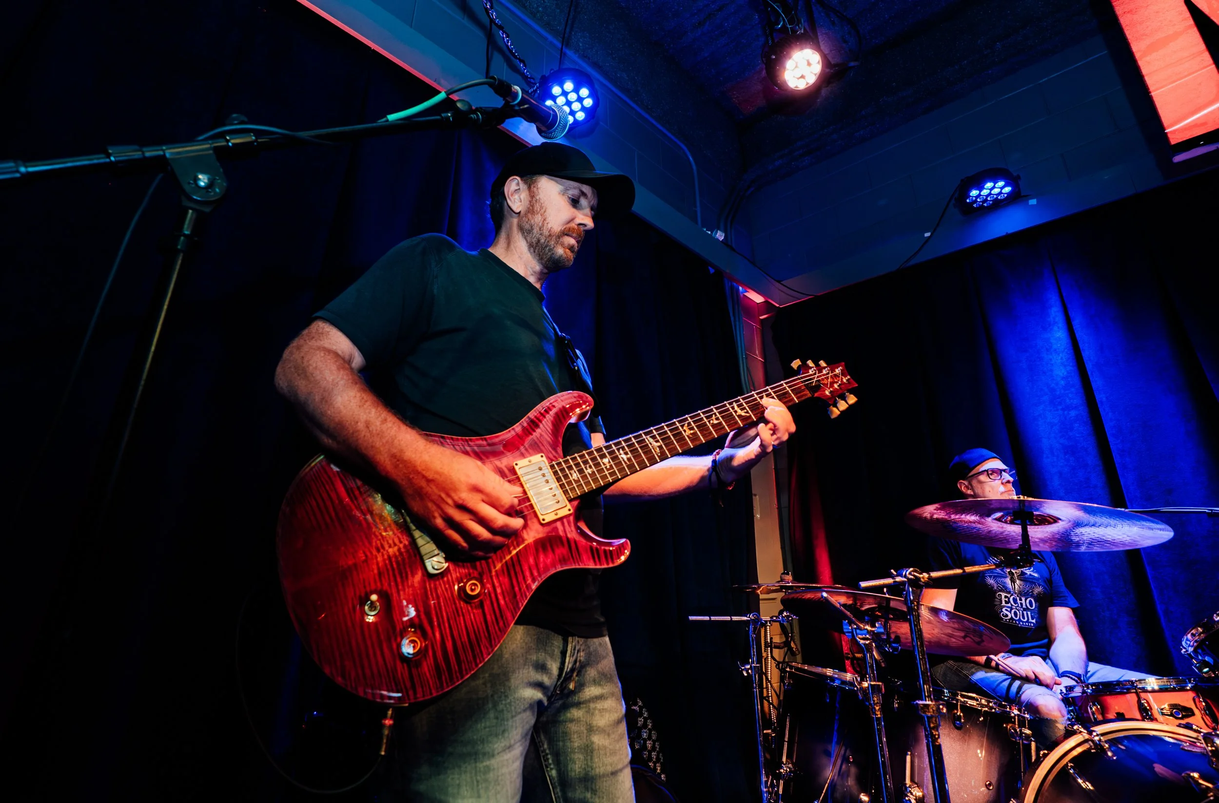Musicians performing on stage with a guitarist in the foreground and a drummer in the background, illuminated by colorful stage lights.