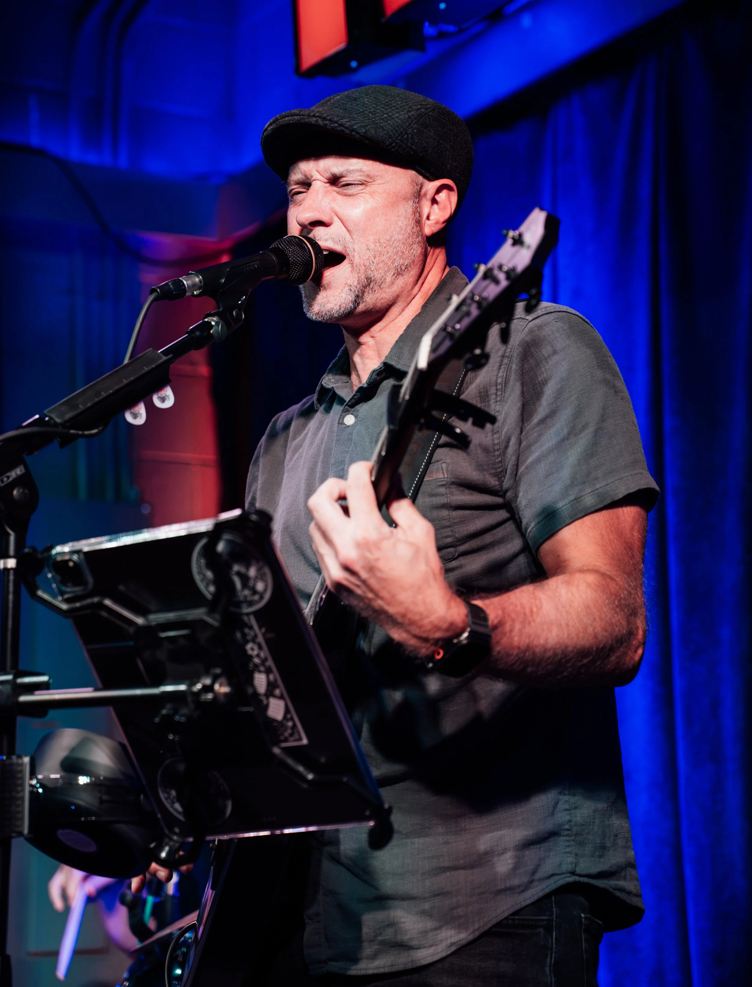 A man singing into a microphone while playing guitar on stage. He is wearing a cap and a gray shirt, with closed eyes and visible emotion. The background has blue curtains and a large screen.