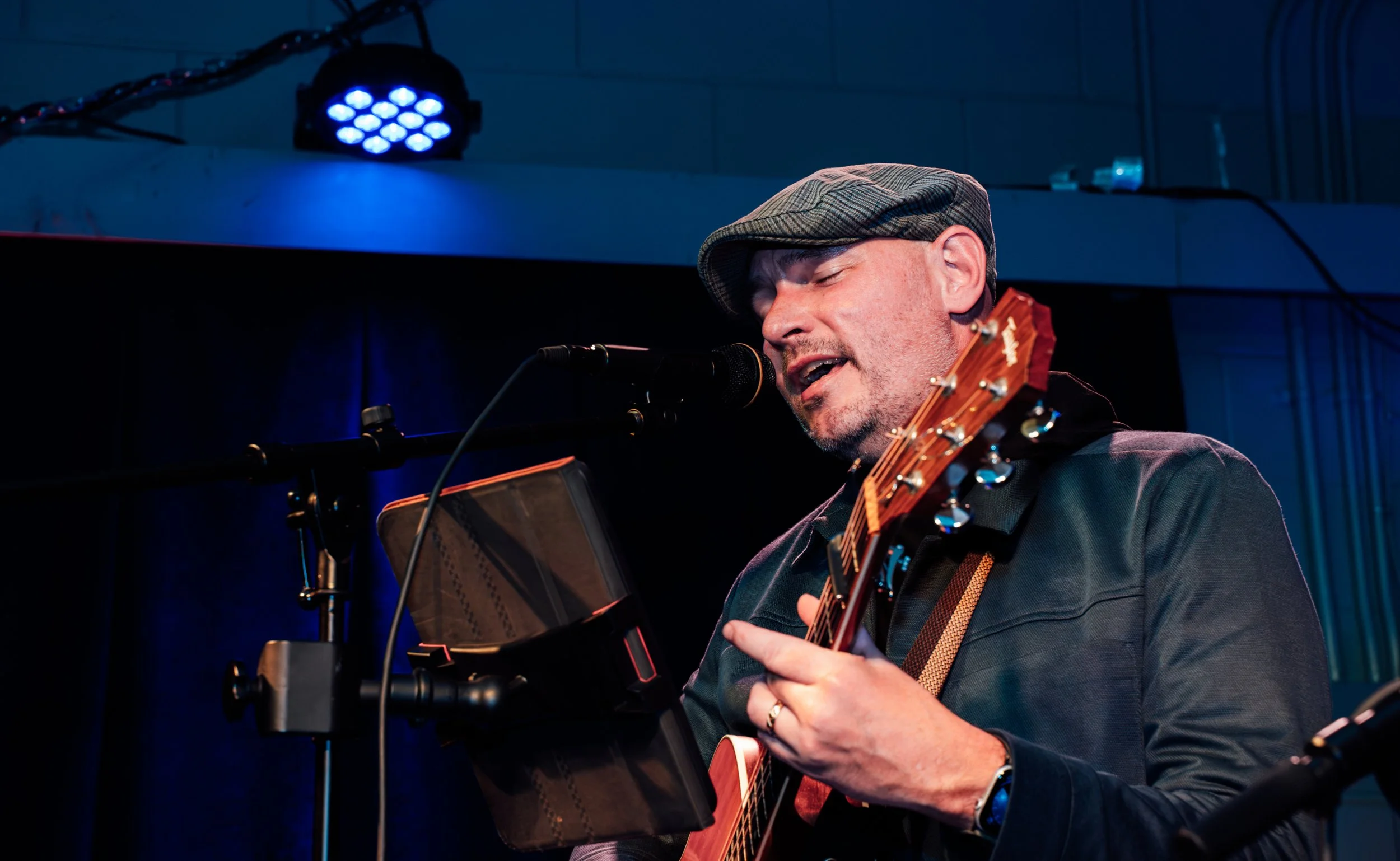 A man wearing a plaid flat cap and a dark jacket performs singing and playing an acoustic guitar on stage, with a blue spotlight overhead.