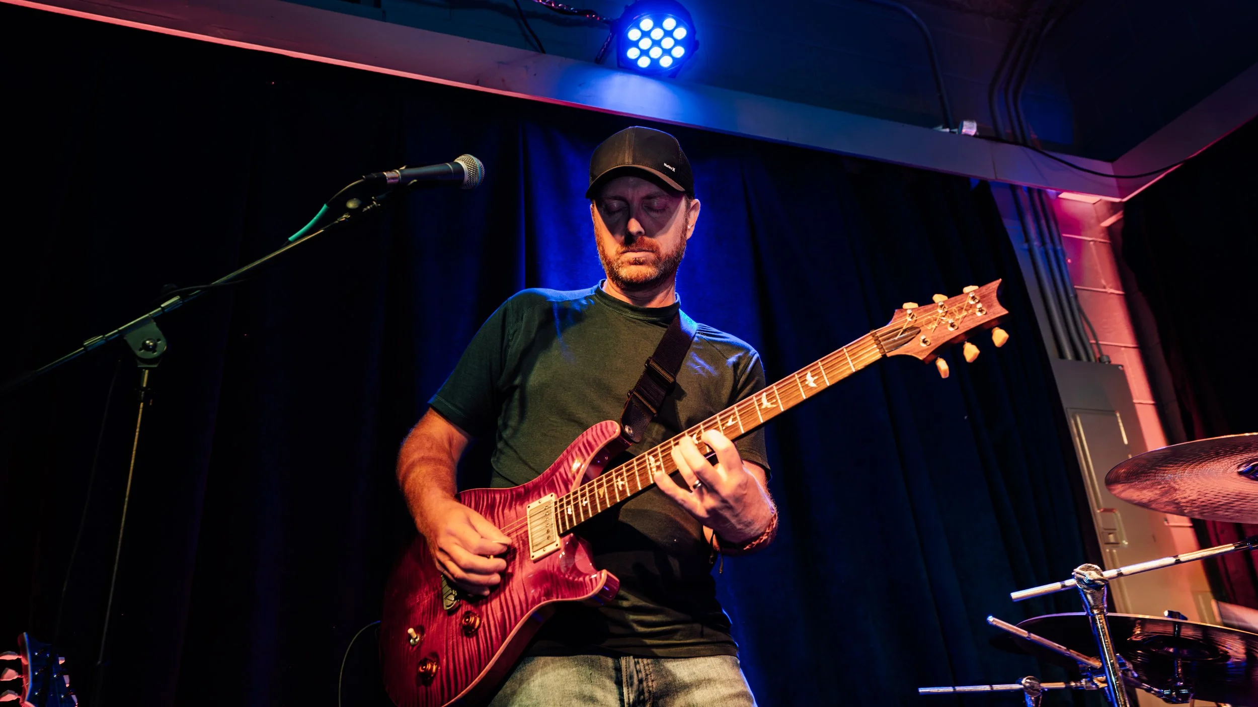 A man wearing a black cap and black t-shirt playing a red electric guitar on stage, with a microphone in front of him and blue stage lighting overhead.