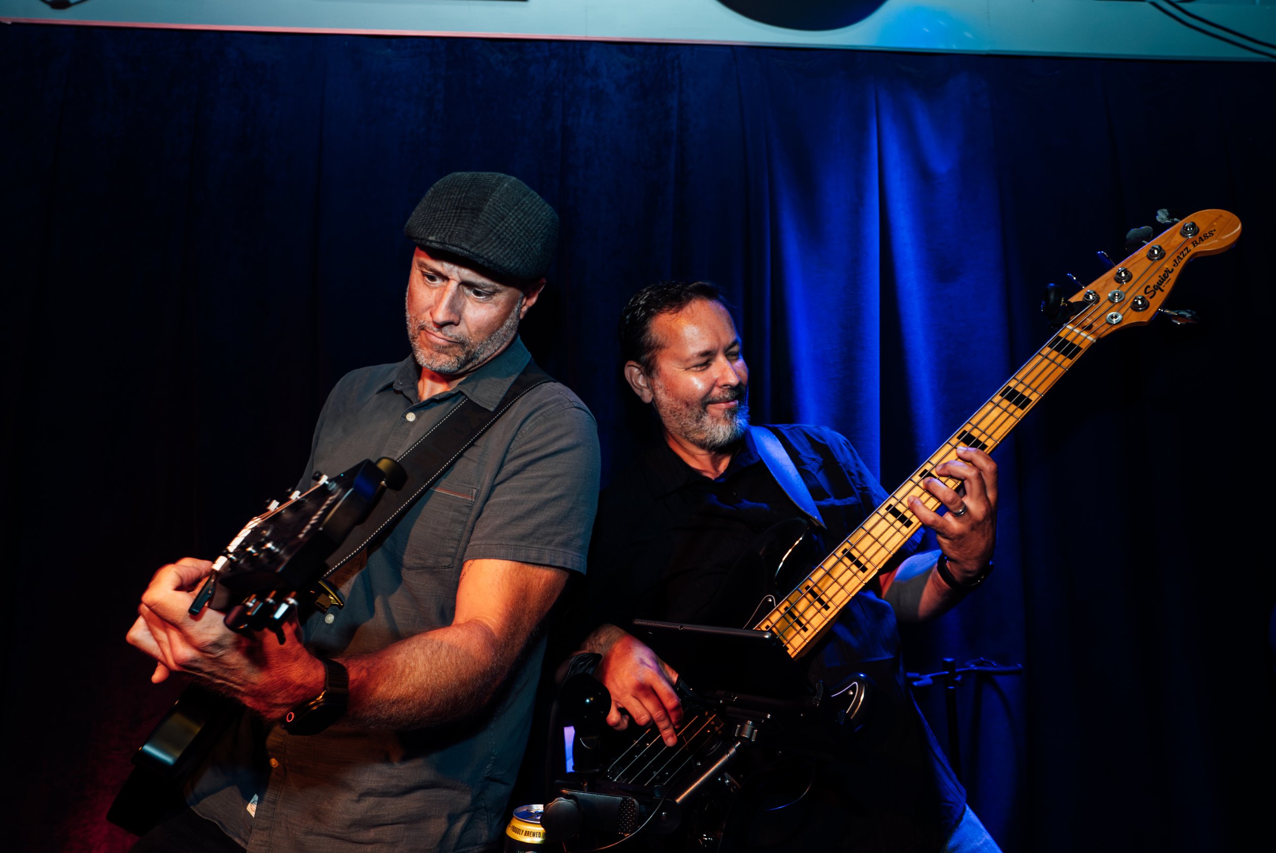 Two men playing guitars on stage, one with a flat cap and the other with dark hair, in front of a dark blue curtain.