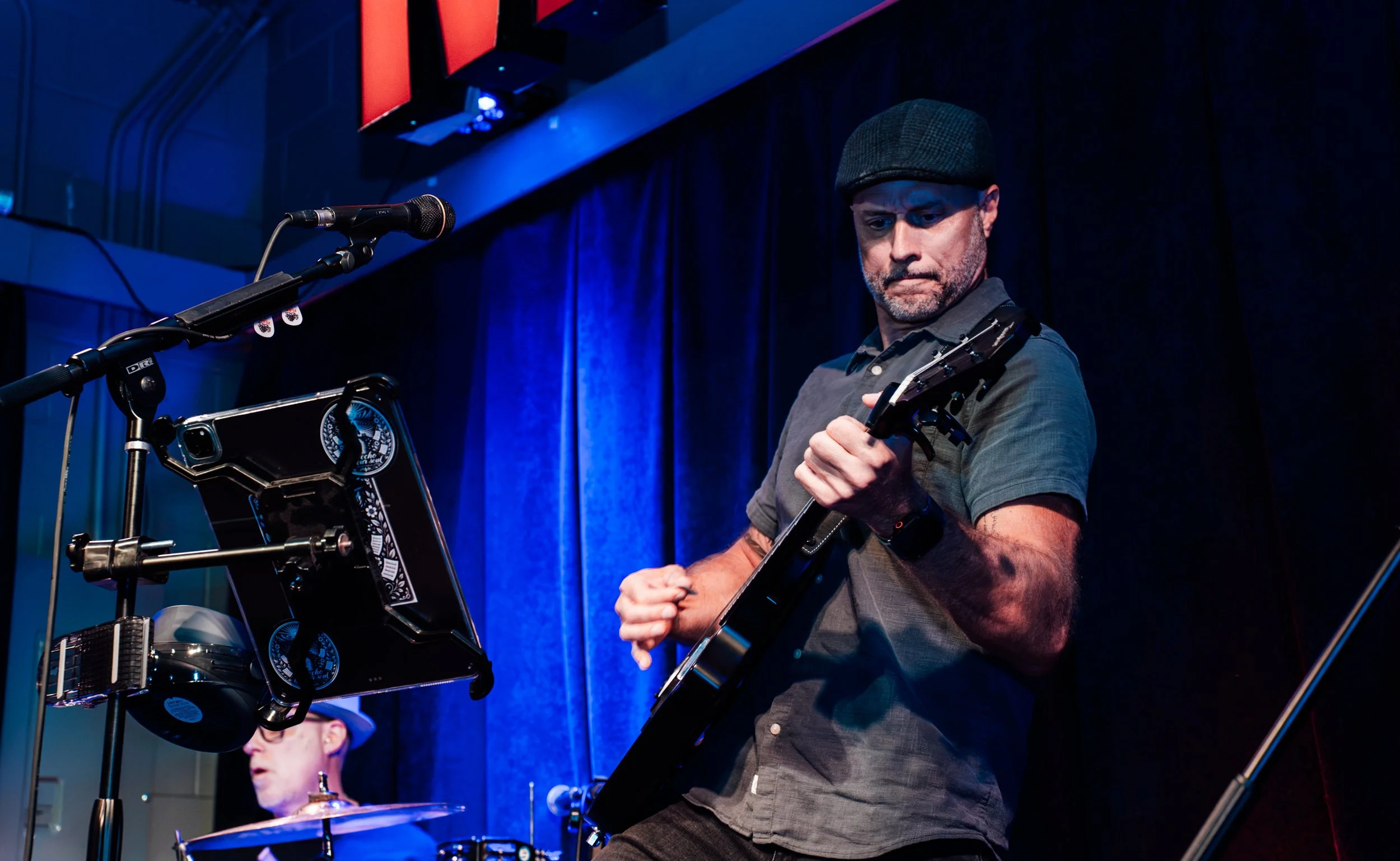 A man playing an electric guitar performing on stage with a blue curtain background, accompanied by drummer in a music venue.