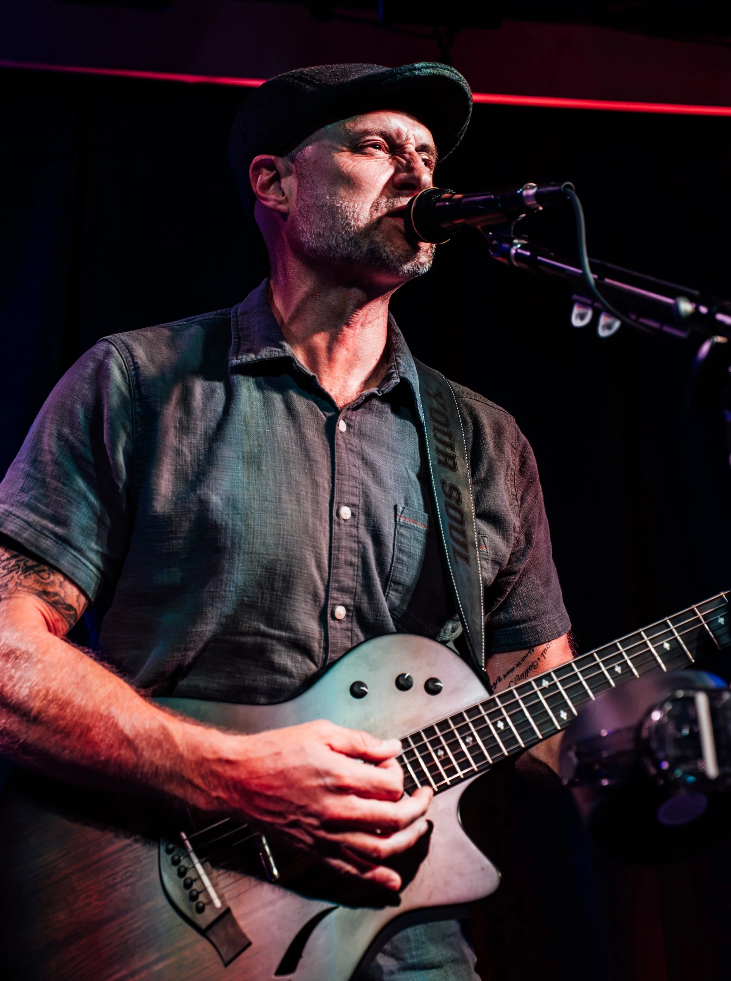 A man with a beard and tattoos, wearing a black cap and a gray shirt, is singing into a microphone while playing an electric guitar in a dimly lit setting.