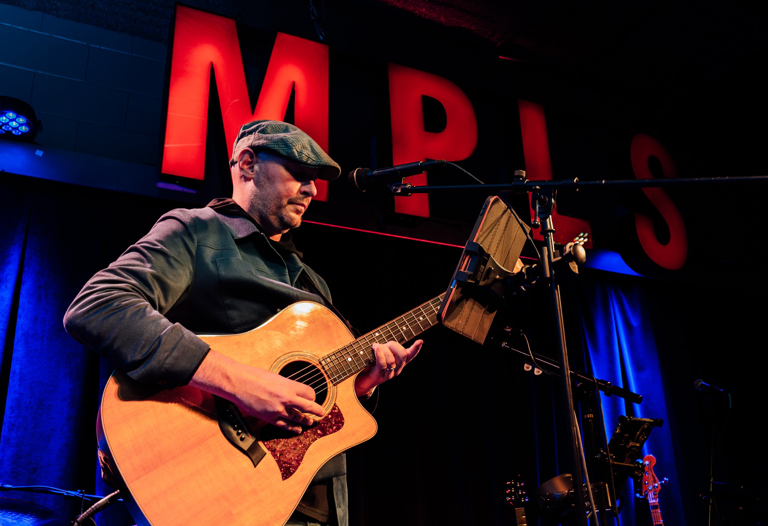 A man wearing a cap and glasses playing an acoustic guitar on stage with red and blue lighting, in front of a large red sign that says 'MIDI'.