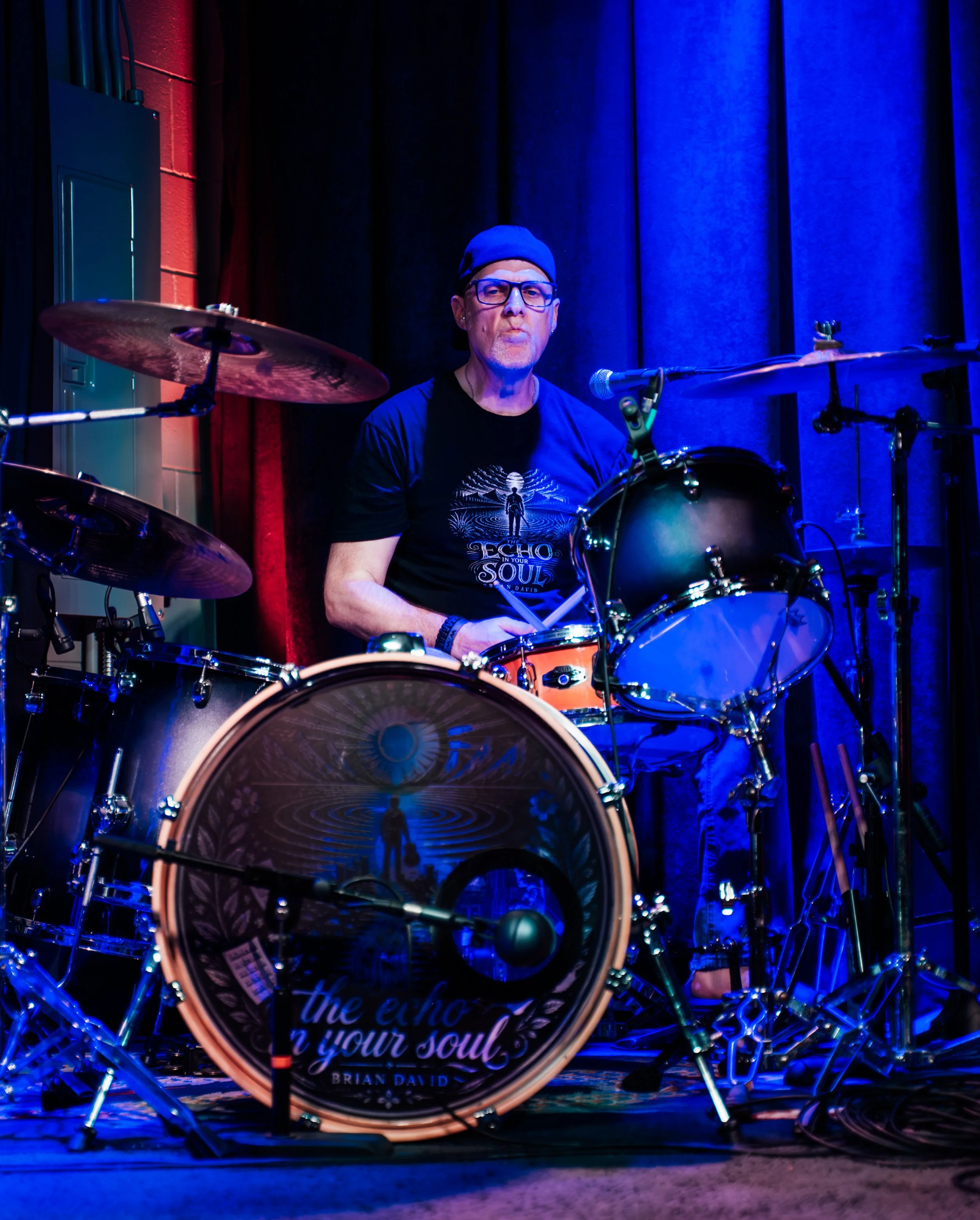 A man with glasses, a beanie, and a black t-shirt sitting behind a drum set illuminated by blue stage lighting.