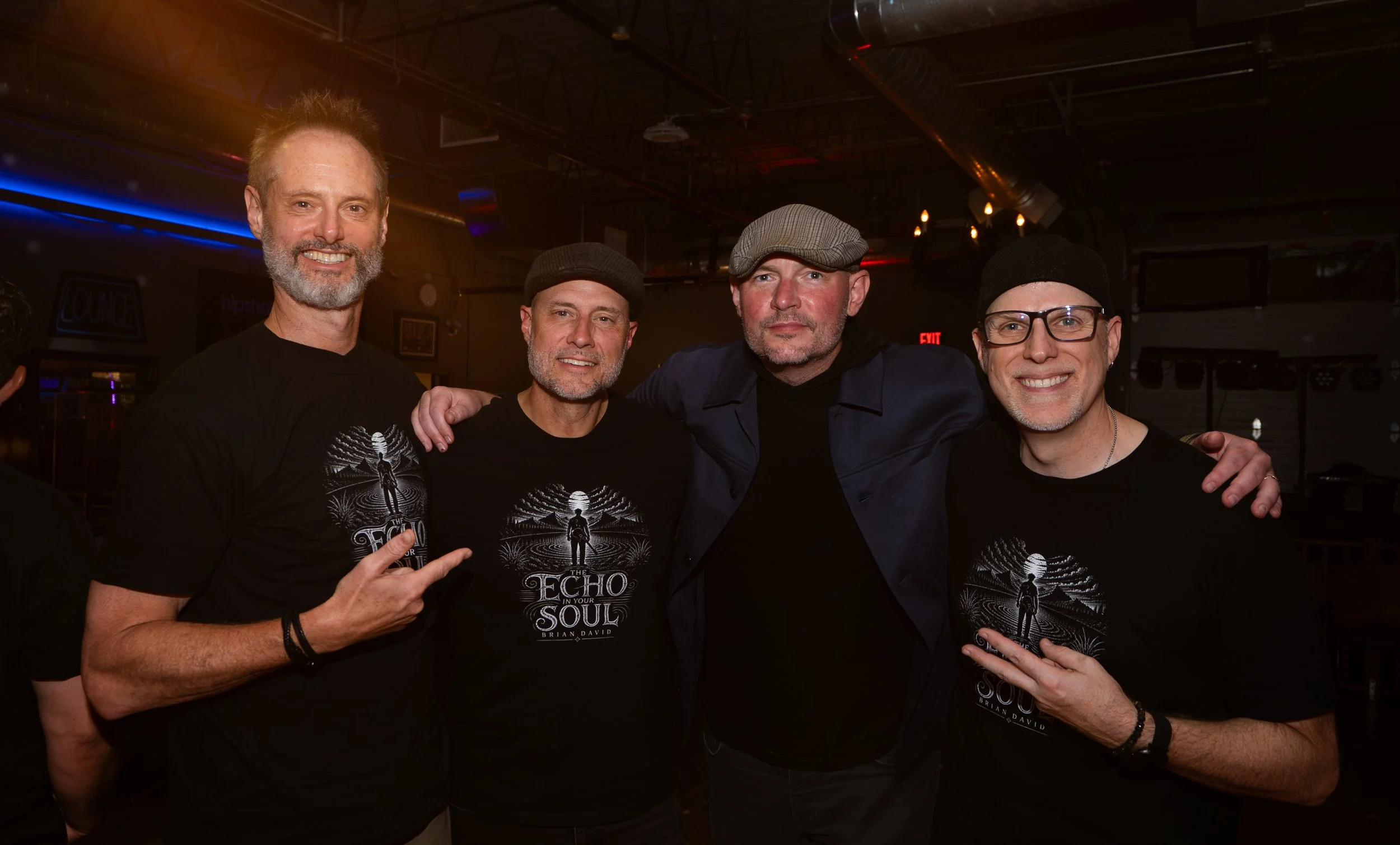 Four men with gray beards and wearing black and gray clothing, standing together indoors, posing for the camera. The background shows a dimly lit entertainment venue with a sign that reads 'LOUNGE.'