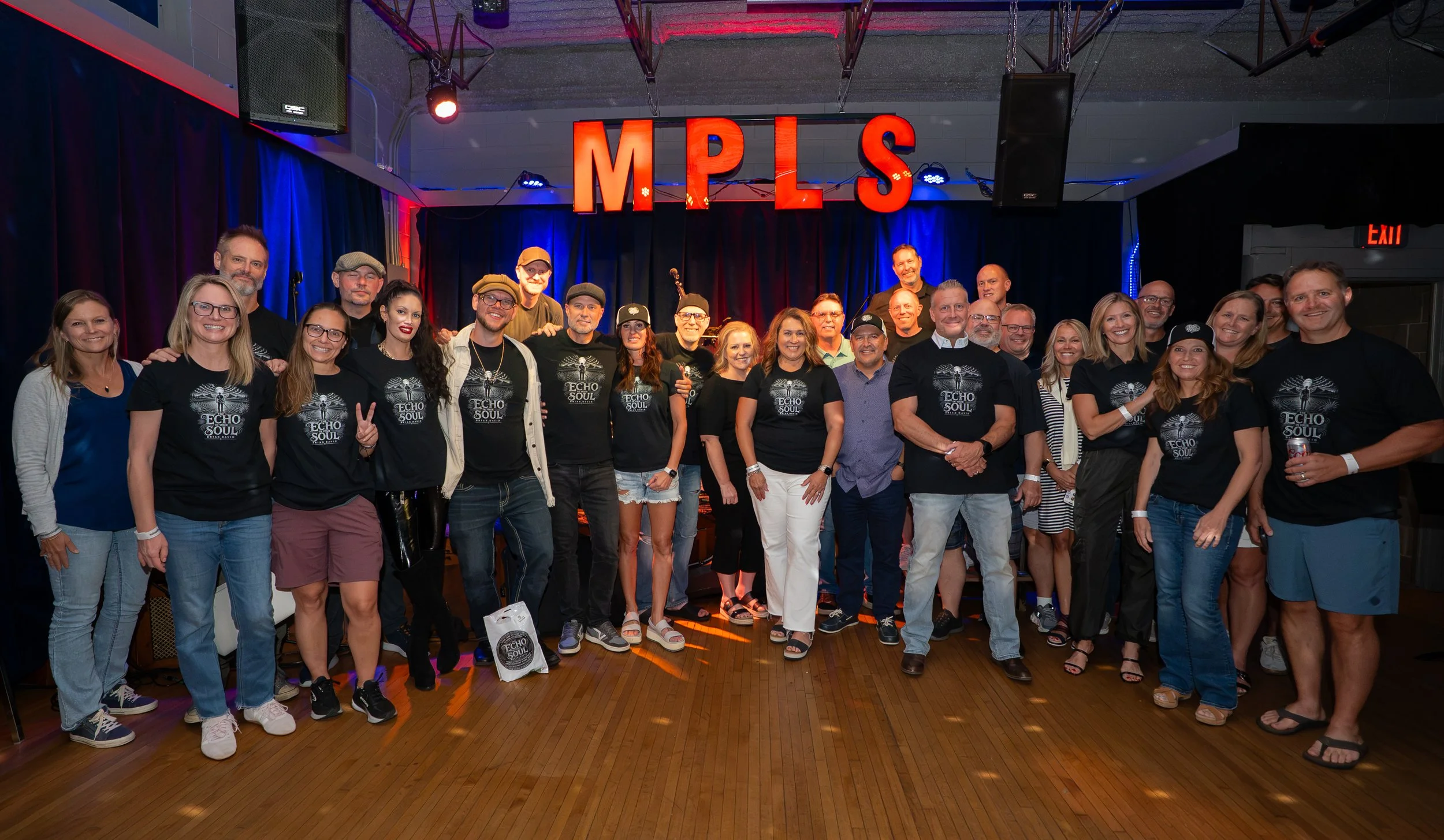 Group of people posing on stage with 'MPLS' sign, some wearing 'Echo Soul' shirts, in a venue with blue and red lighting.