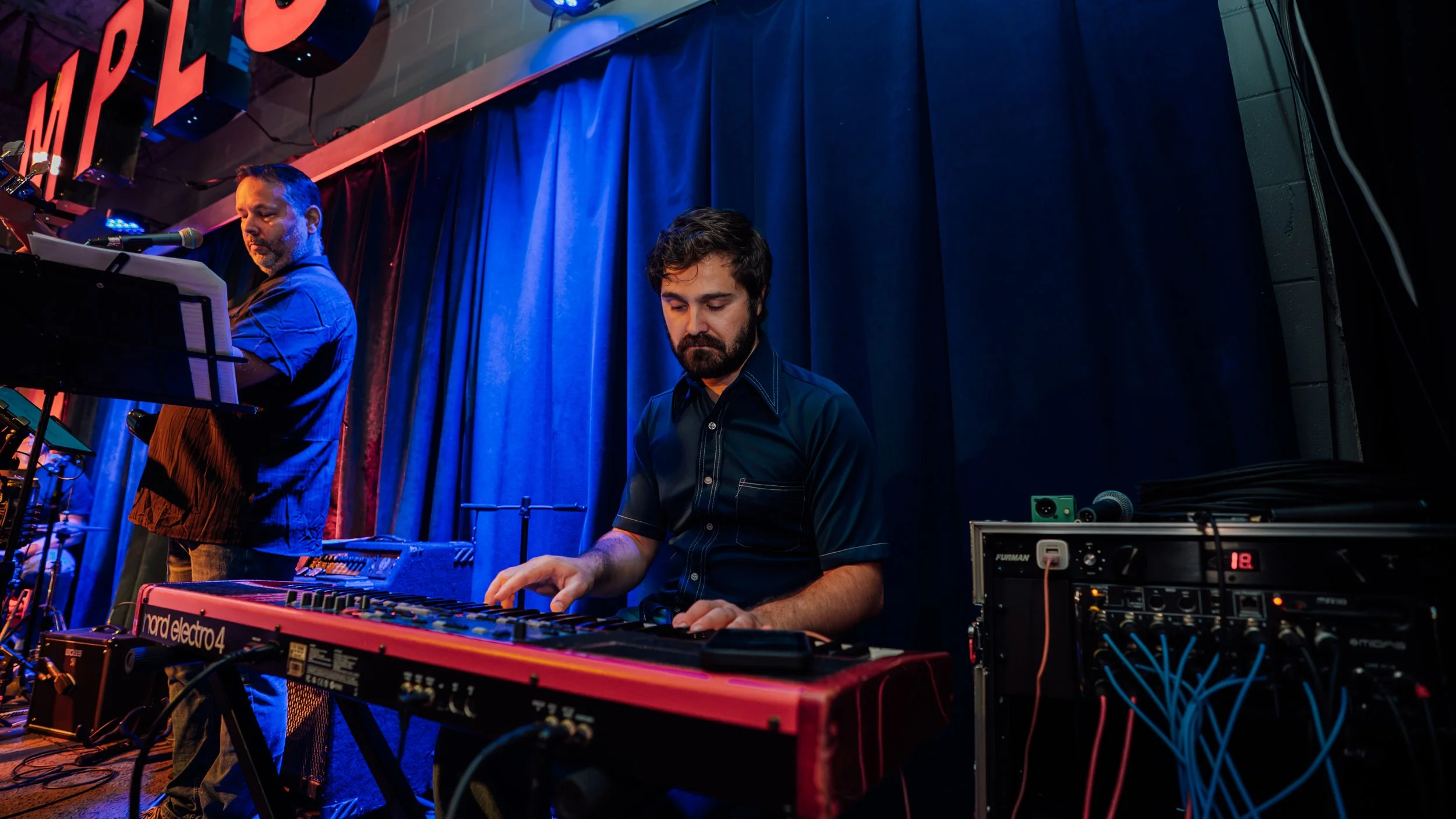 A musician with a beard and dark shirt playing a keyboard on stage, with another person behind him also performing. The stage has blue curtains and various electronic equipment and cables.