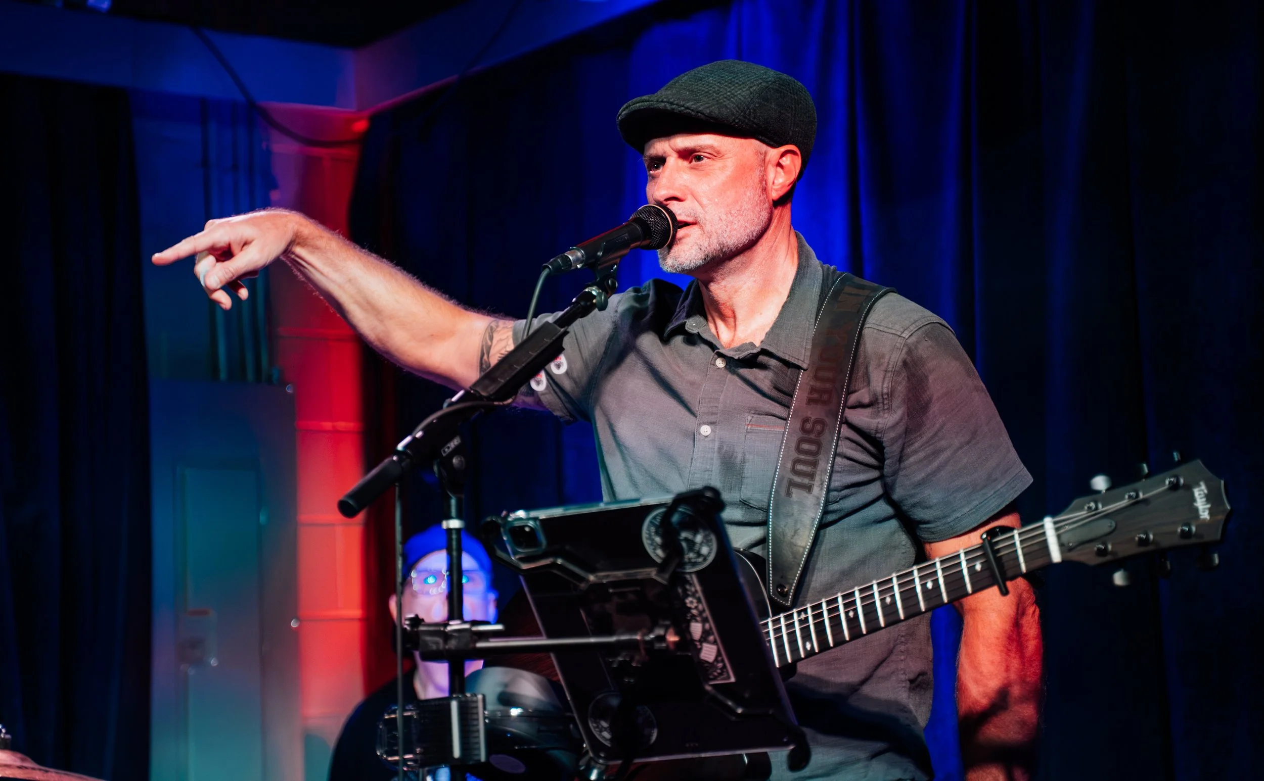 A man performing on stage, singing into a microphone, wearing a black flat cap, gray short-sleeve shirt, and playing an acoustic guitar. The background has blue and red lighting with curtains.