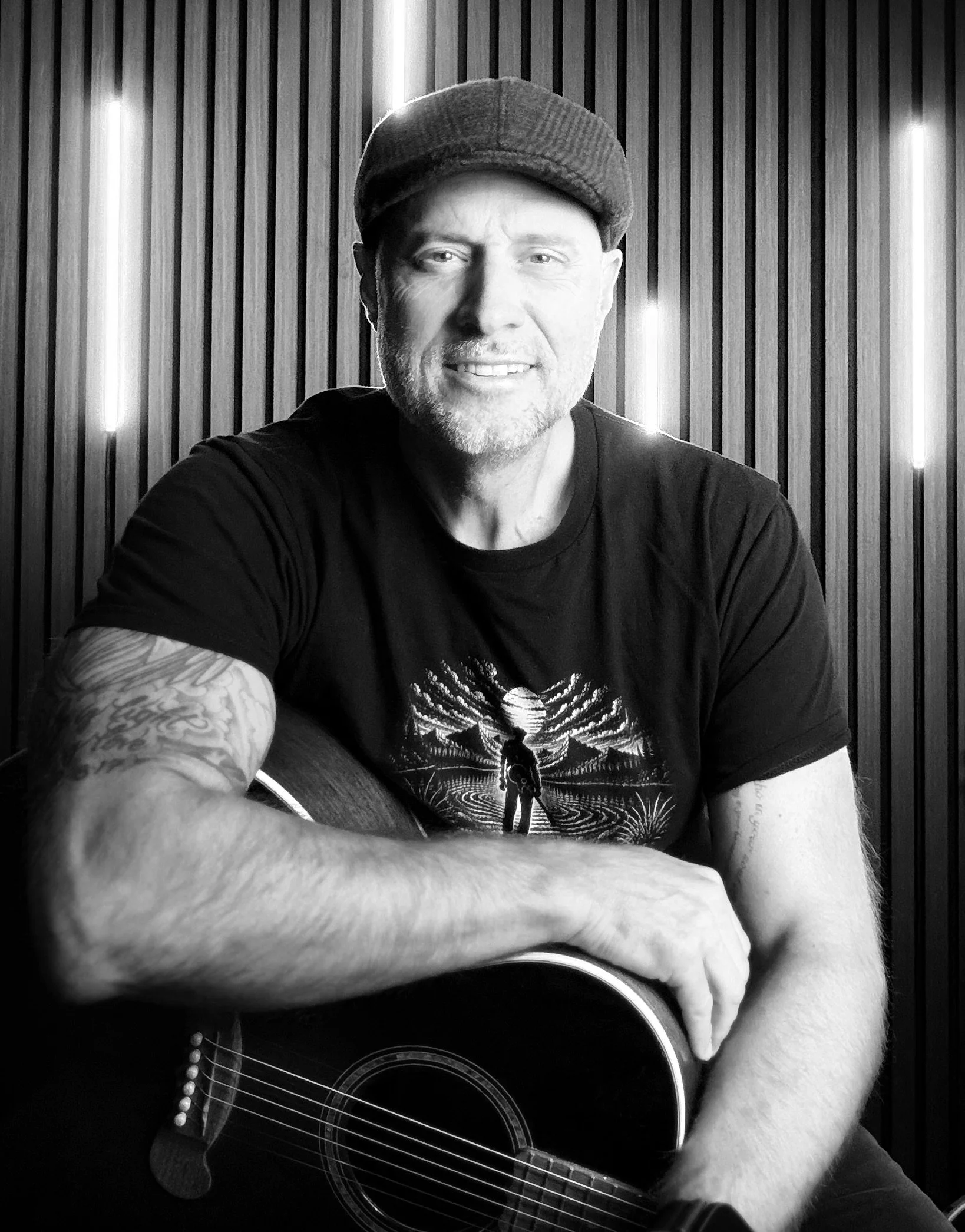 Black and white photo of a man wearing a cap and a T-shirt with a skeleton design, playing an acoustic guitar.