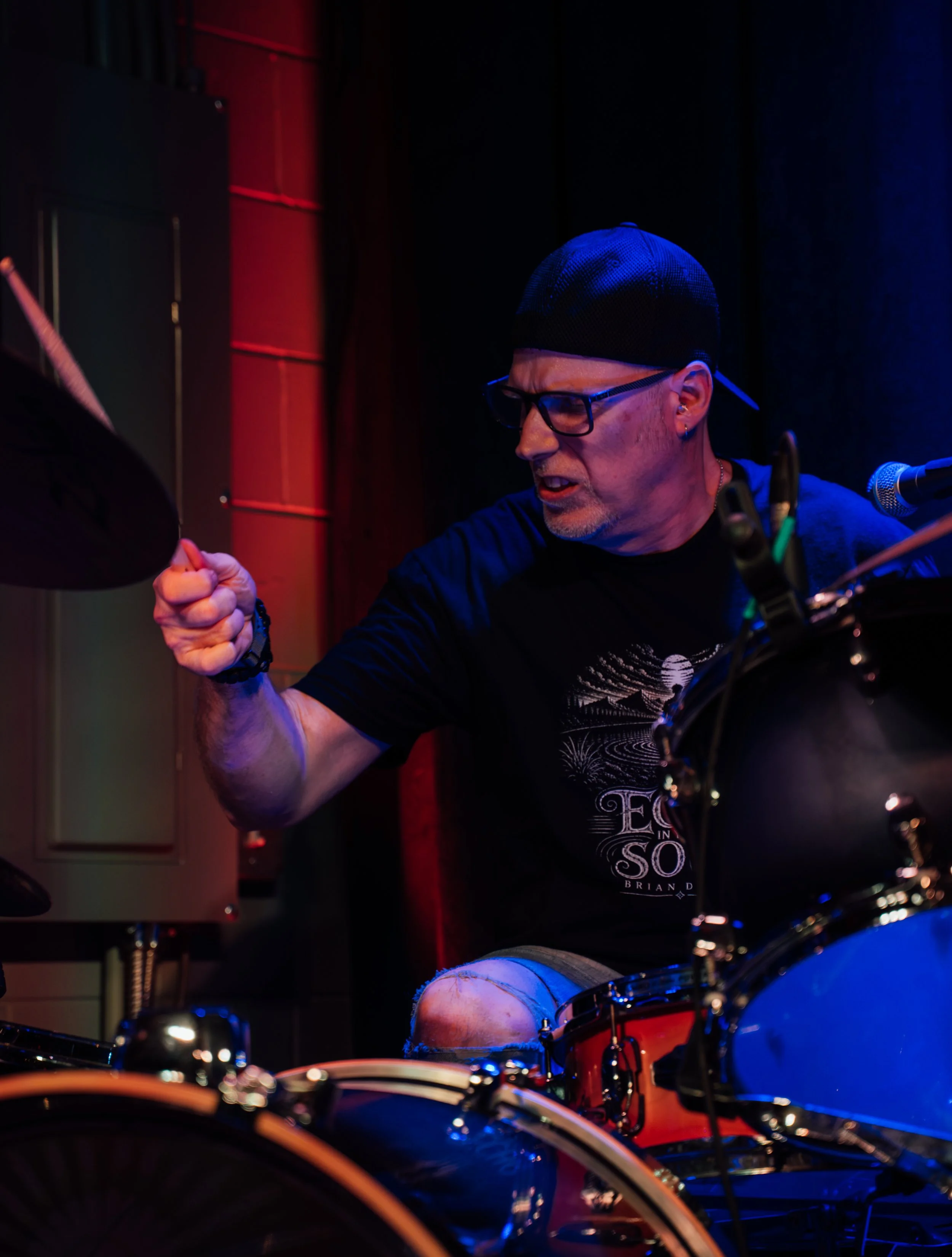 A man wearing glasses, a black cap, and a T-shirt with a design, is playing drums or percussion in a dimly lit setting with colorful stage lighting.