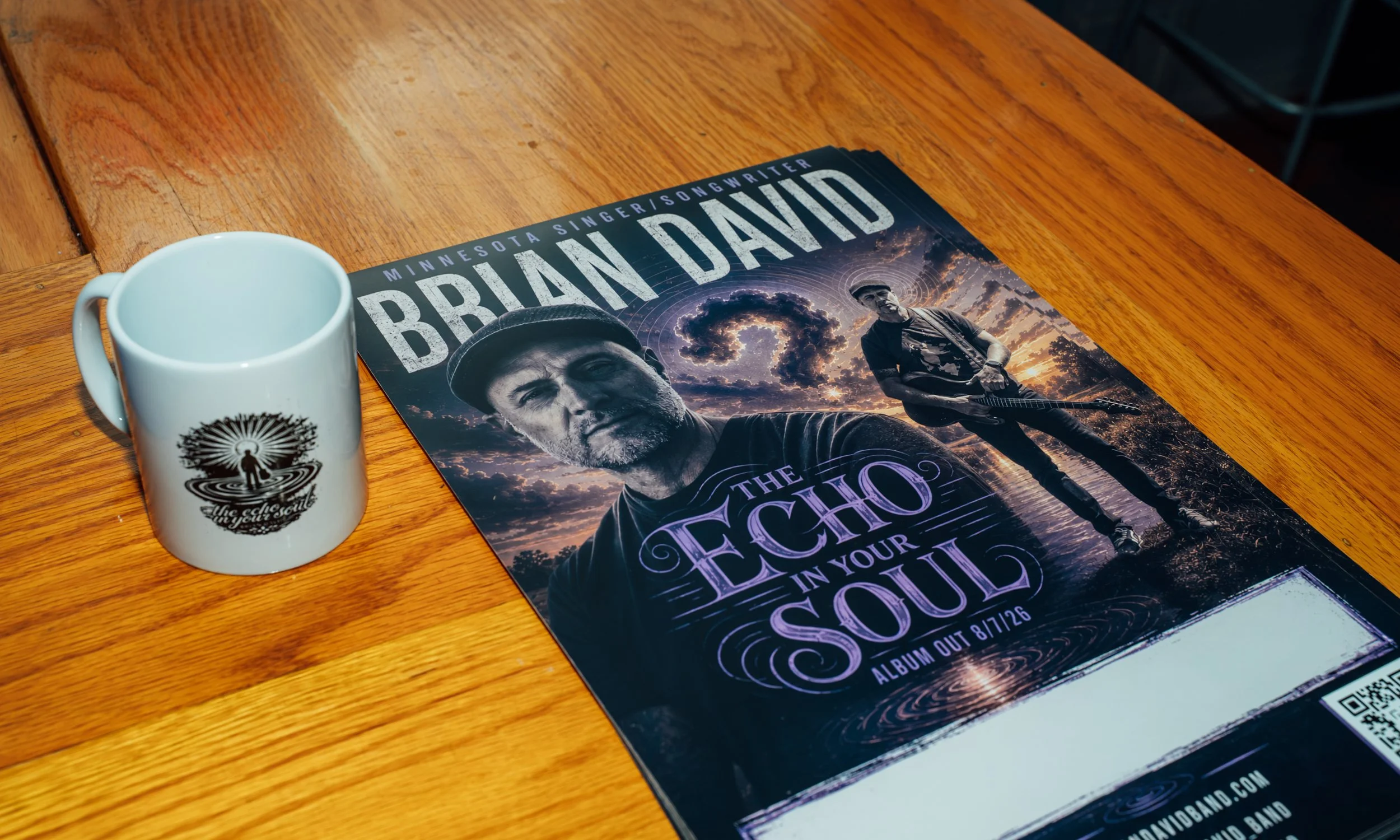 A white mug with black spiral designs and a black silhouette of a person on it sits on a wooden table next to a concert flyer for Brian David and The Echoes in Your Soul. The flyer features a man with a beard, wearing a hat, holding a guitar, with a 