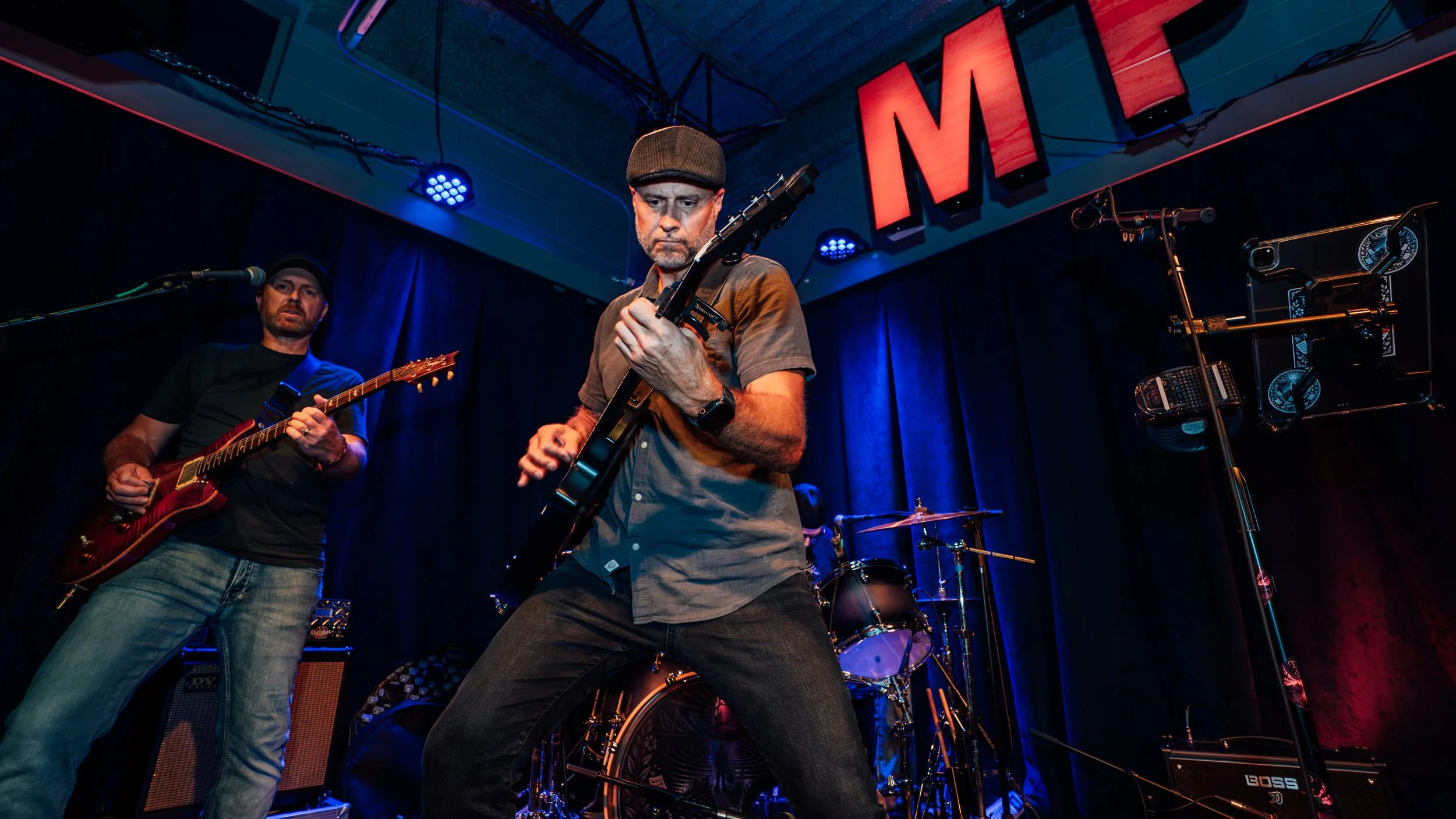 Two musicians performing on stage with a blue curtain backdrop. One is playing an electric guitar and the other is playing a bass guitar. Large red illuminated letters spell 'MI' above them. Stage lights are visible, and there are musical equipment a