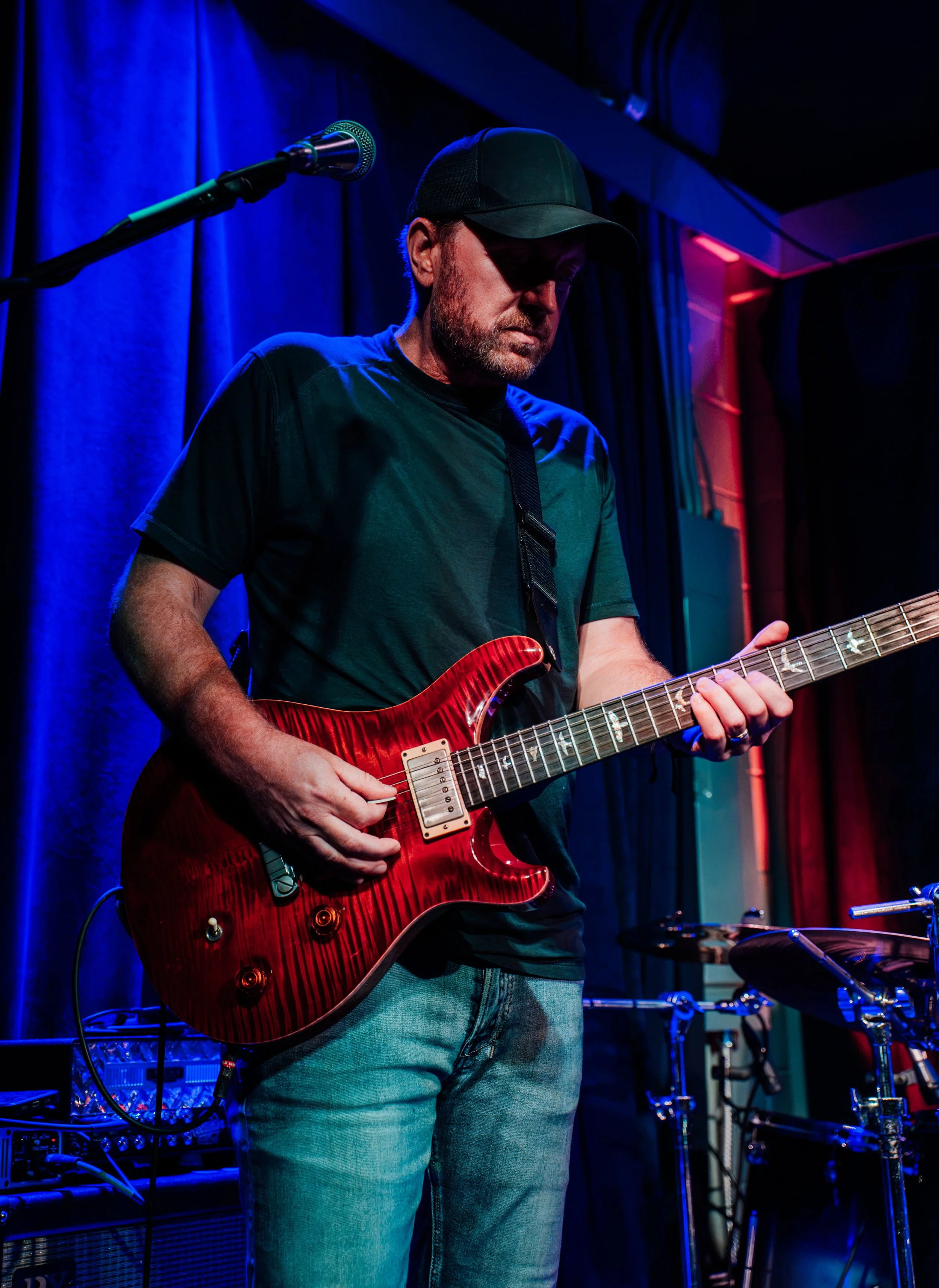 A man playing an electric guitar on stage with blue and red stage lighting, head slightly bowed, wearing a black cap, a dark green t-shirt, and jeans.