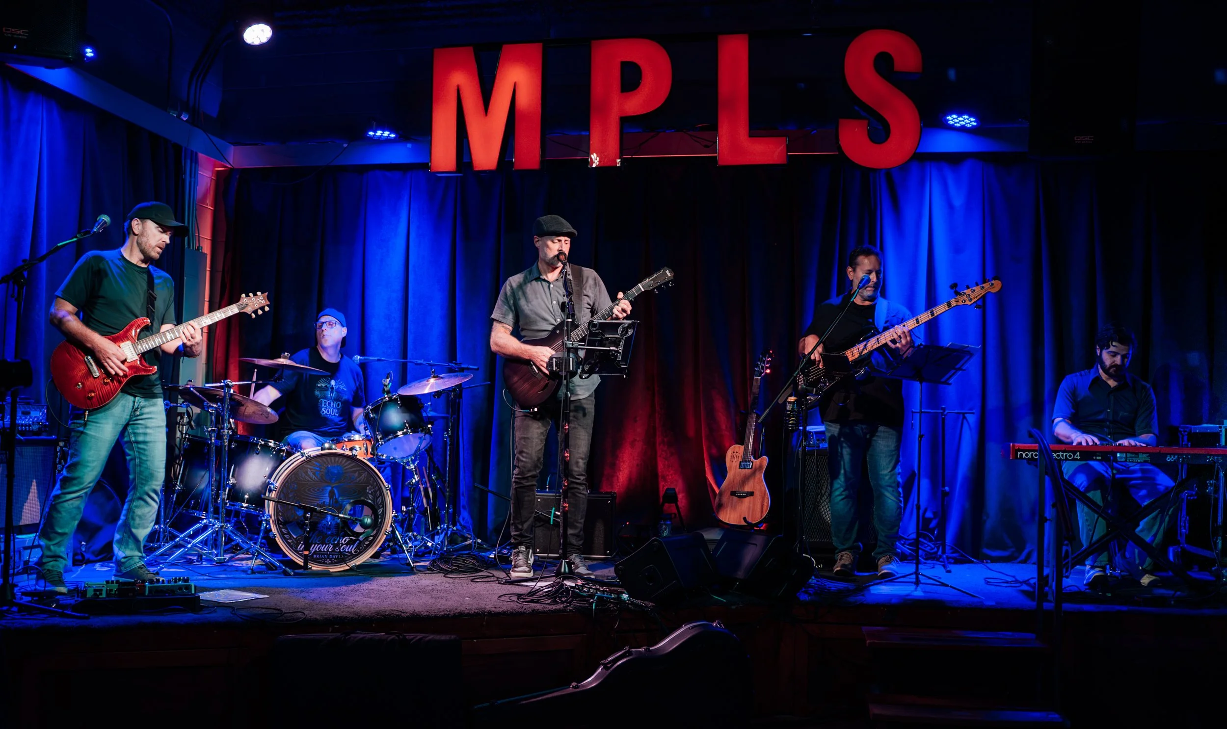 A band of five musicians performing on stage at MPIS, with a backdrop of red and blue curtains, blue stage lighting, and musical instruments including guitars, drums, and a keyboard.