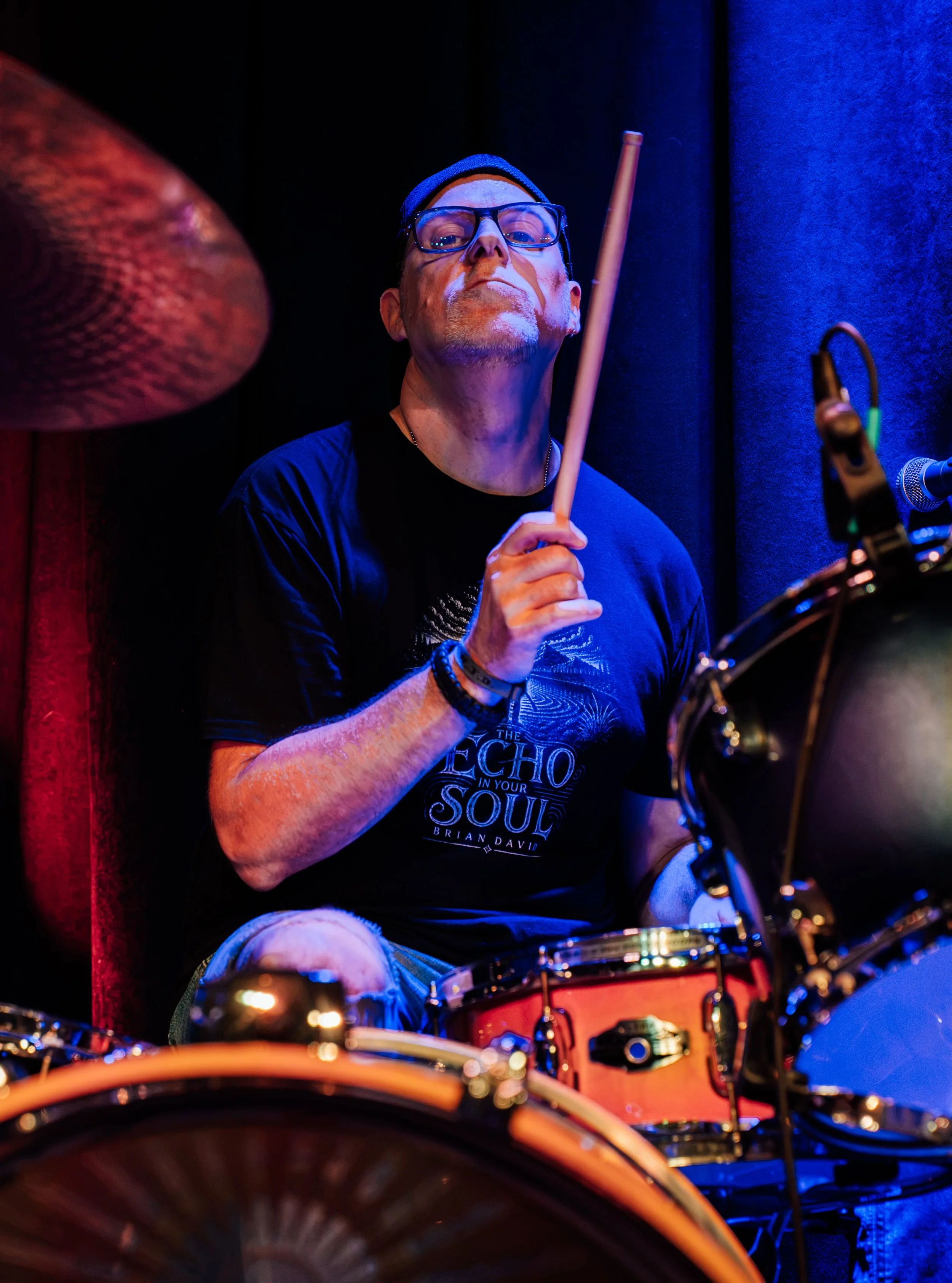 A man wearing glasses, a black beanie, and a black t-shirt with the text 'The Echo in Your Soul' is playing drums on a stage with blue and red lighting.