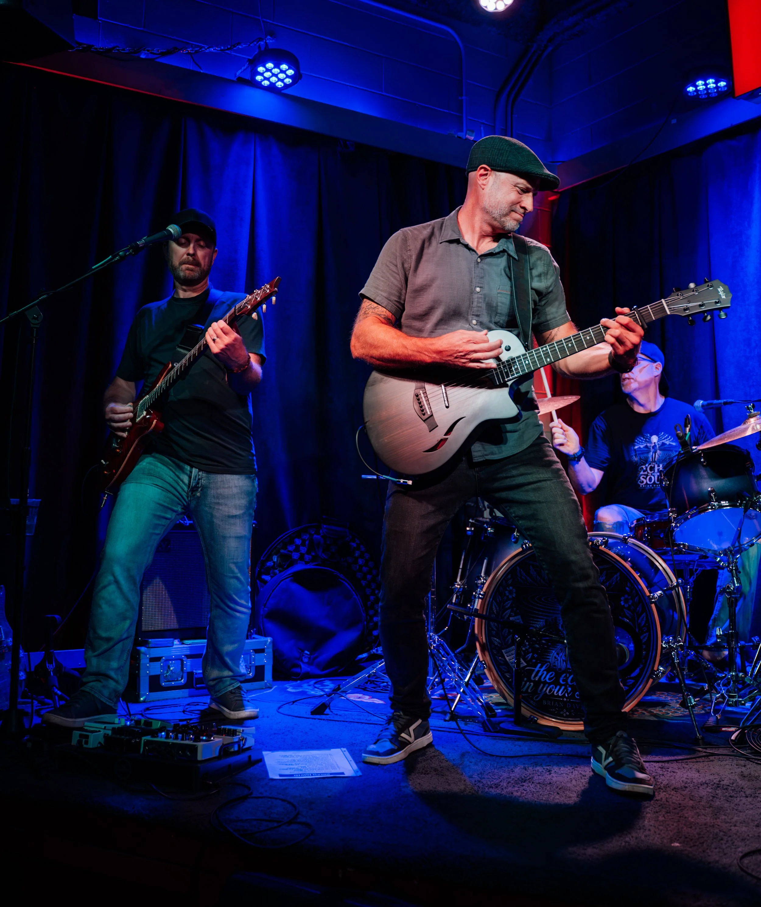 Musicians performing on stage under blue and red lighting, with one person playing an electric guitar and another person playing an acoustic guitar, while a woman in the background plays the drums.