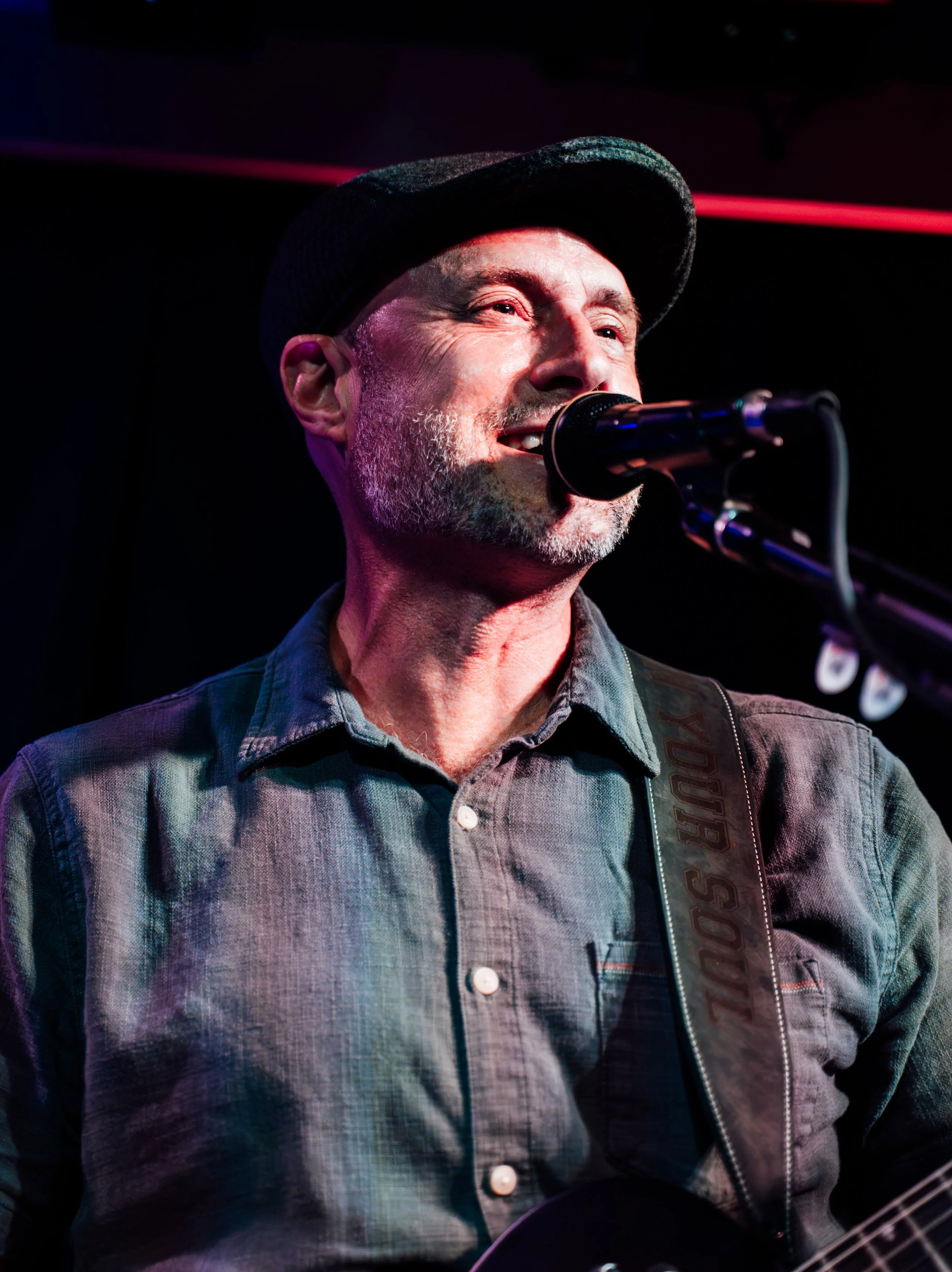 A man with a grey beard and mustache, wearing a dark hat, singing or speaking into a microphone on stage.
