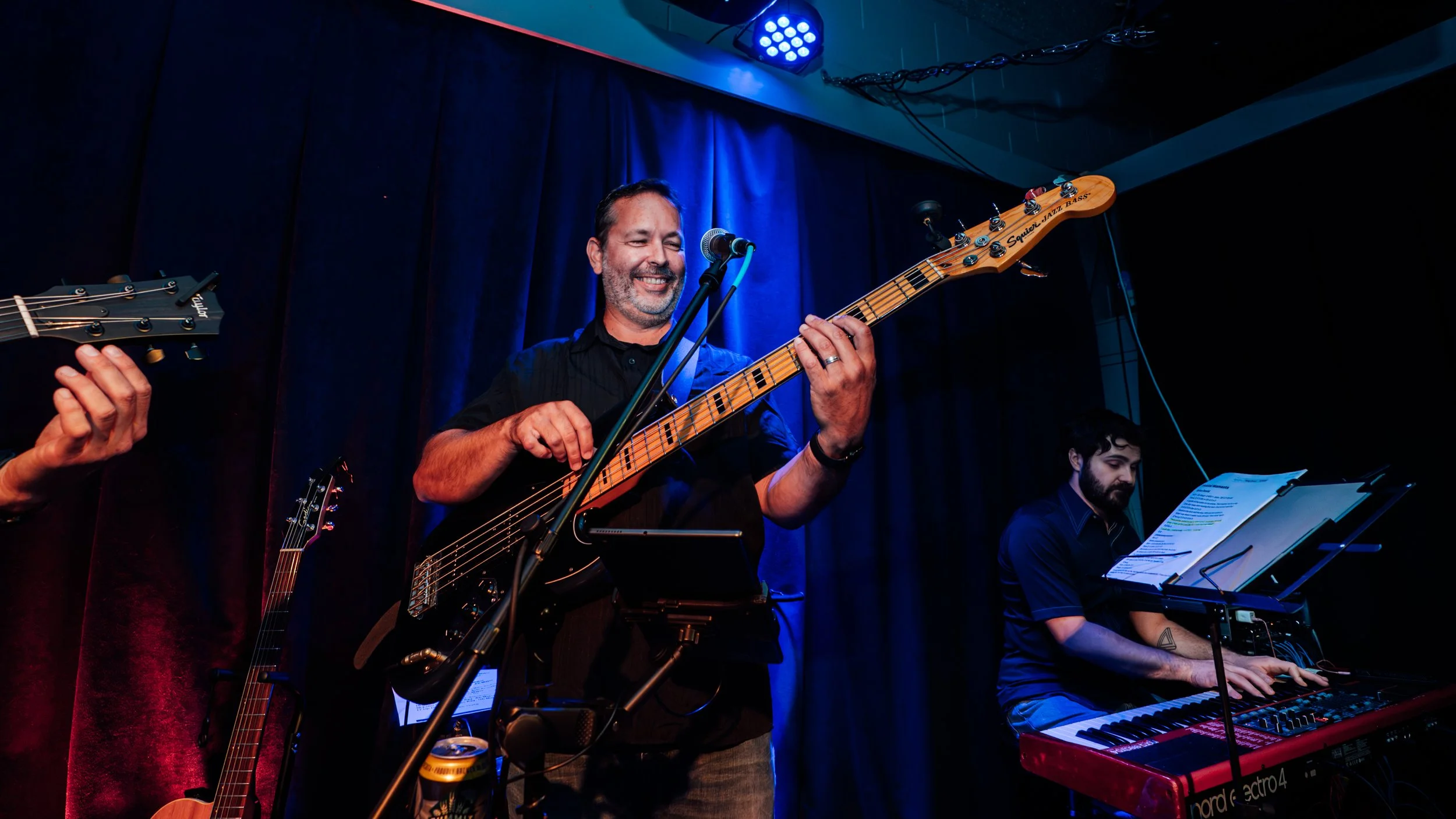 Musicians performing on stage with a man playing an electric bass guitar, another man playing a keyboard, and a part of a guitar visible on the left, against a blue curtain backdrop.