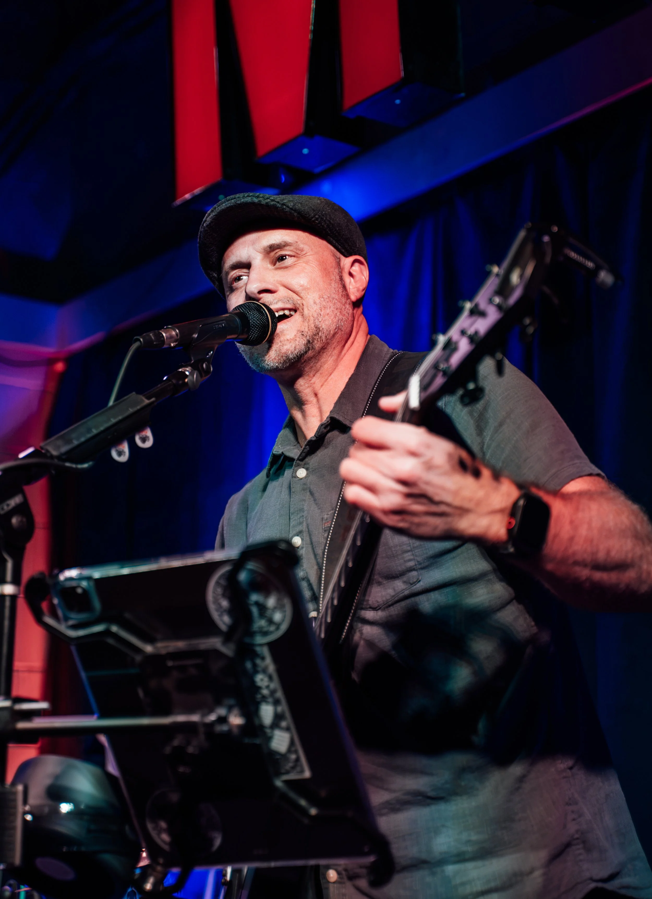 A man wearing a black flat cap playing an electric guitar and singing into a microphone on stage, with a blue and red illuminated background.