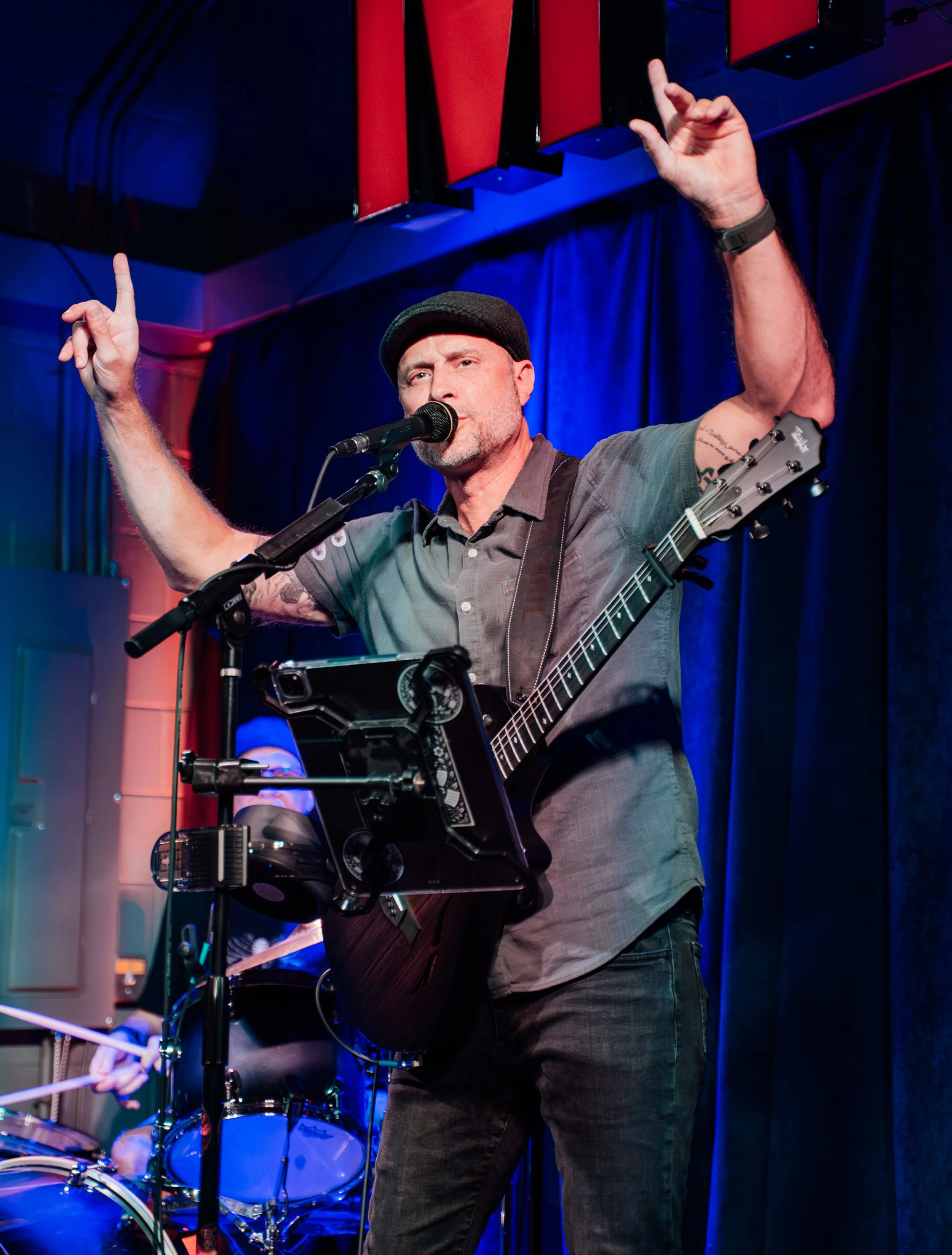 A man wearing a gray shirt and a black cap is singing into a microphone while playing an acoustic guitar during a live performance on stage with a blue and red curtain backdrop.