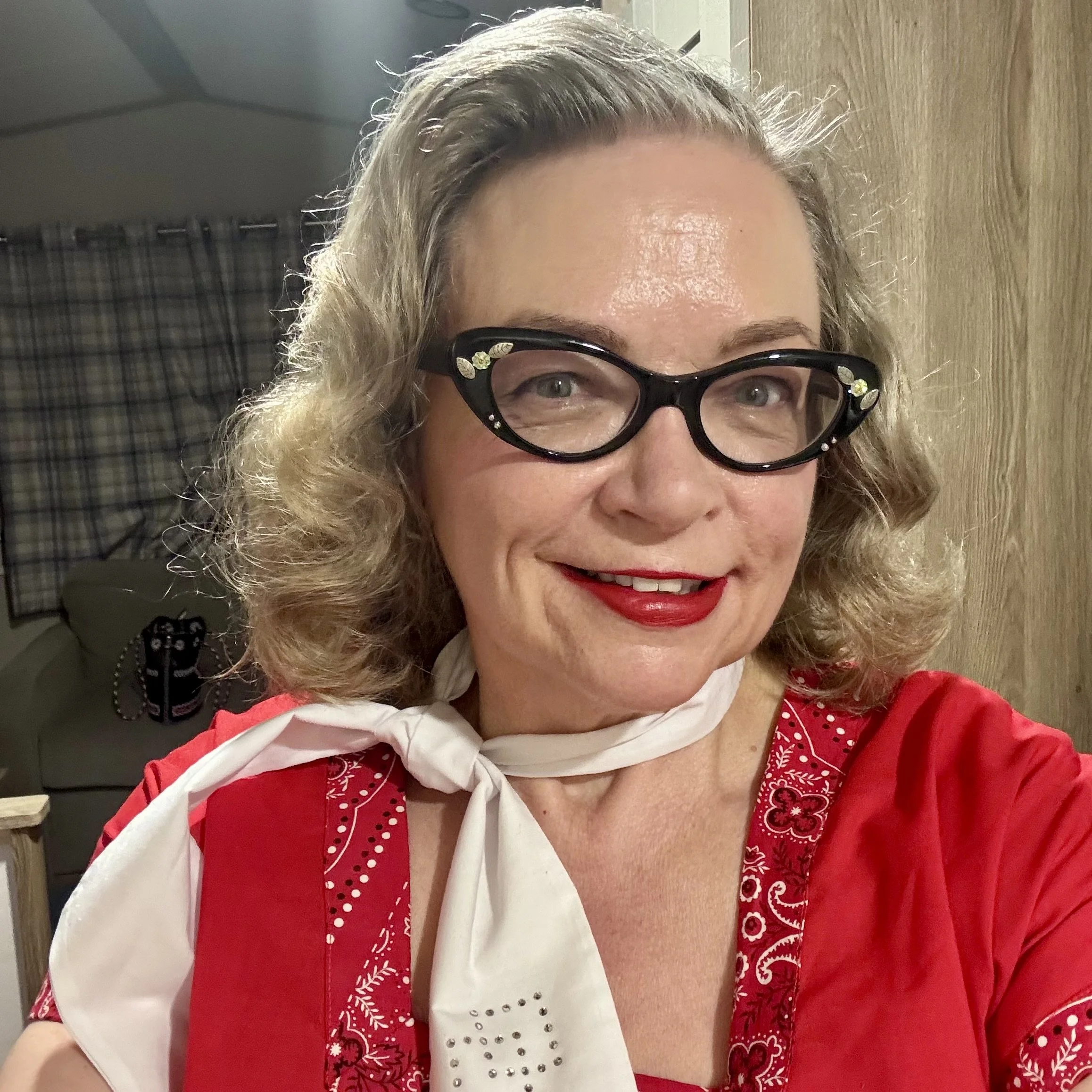A close-up photo of a woman with curly blonde hair, wearing black glasses with decorative embellishments and red lipstick. She is dressed in a red and white outfit with embroidery and a white scarf around her neck. The background shows a plaid curtain and part of a room.
