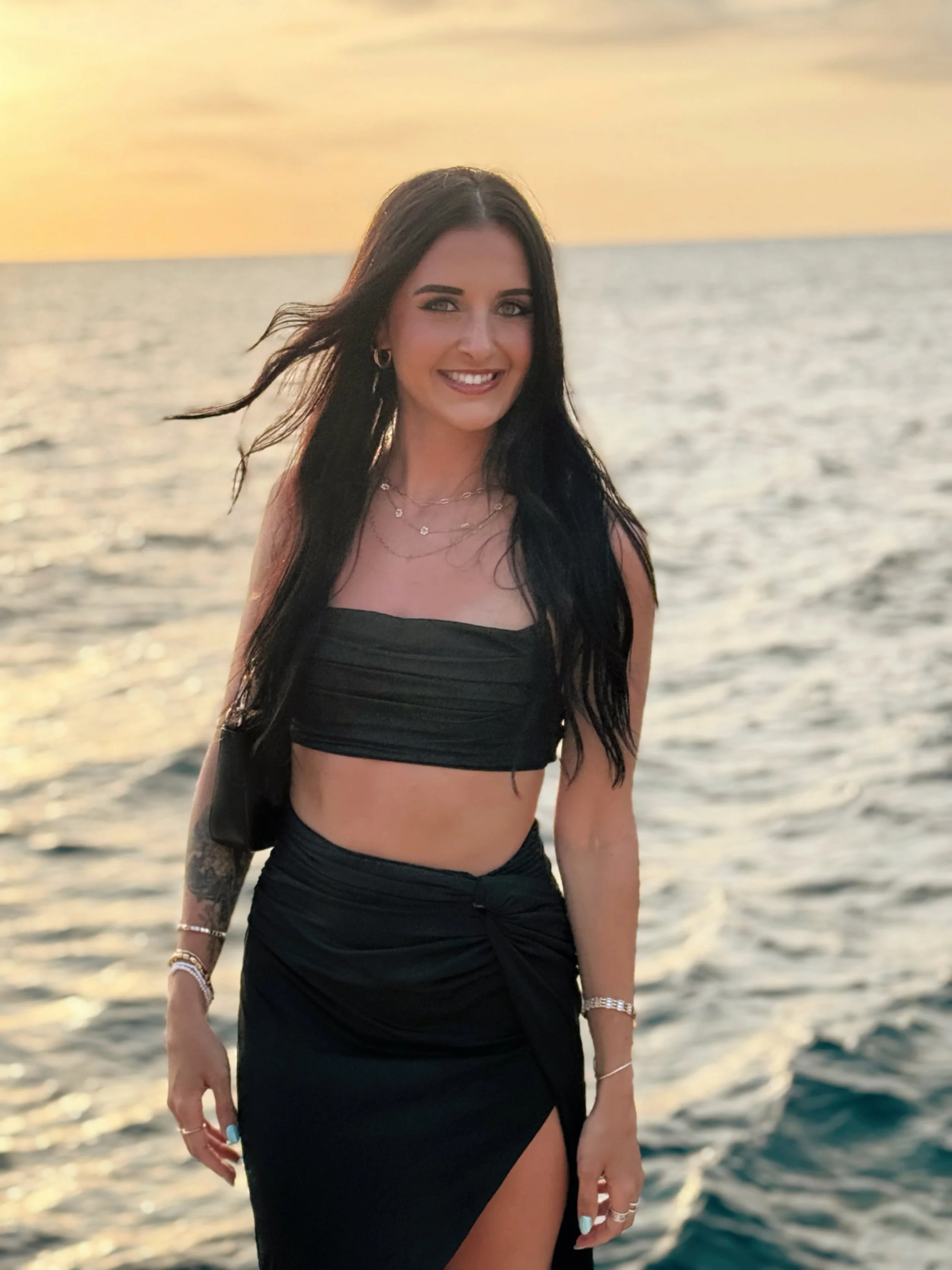 A smiling woman with long dark hair stands by the ocean during sunset, wearing a black strapless crop top and matching skirt with a high slit.