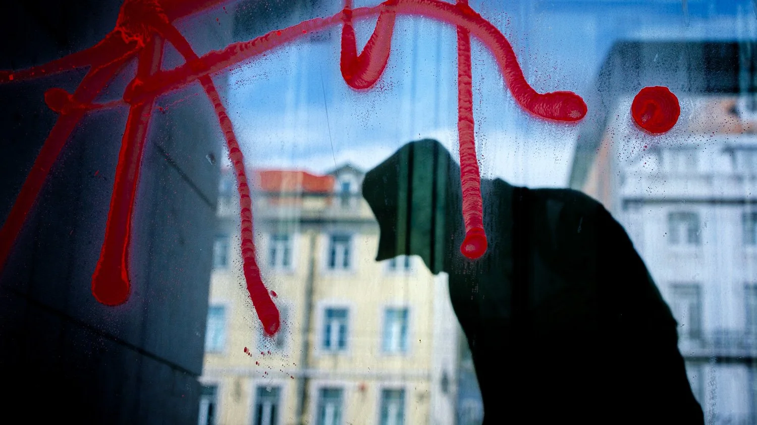 A person cleaning a window with red paint or liquid, creating streaks and drops on the glass, with buildings visible in the background.