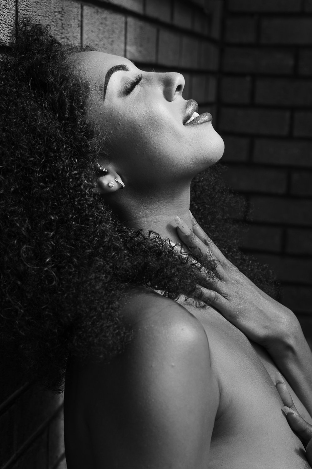 Black and white photo of a woman with curly hair, with eyes closed, head tilted back, and hand on her neck, against a brick wall background.