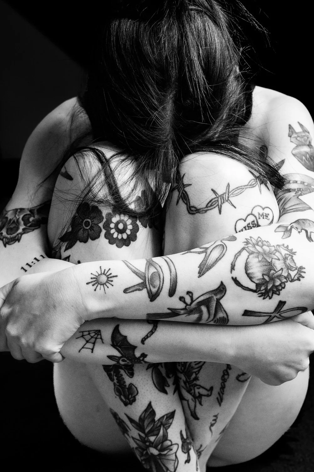 A woman with long dark hair has multiple tattoos on her arms, shoulders, and chest, including flowers, barbed wire, a heart with the words 'Miss Me,' a skull, a dagger, and other designs. She is hugging her knees, and her face is not visible.